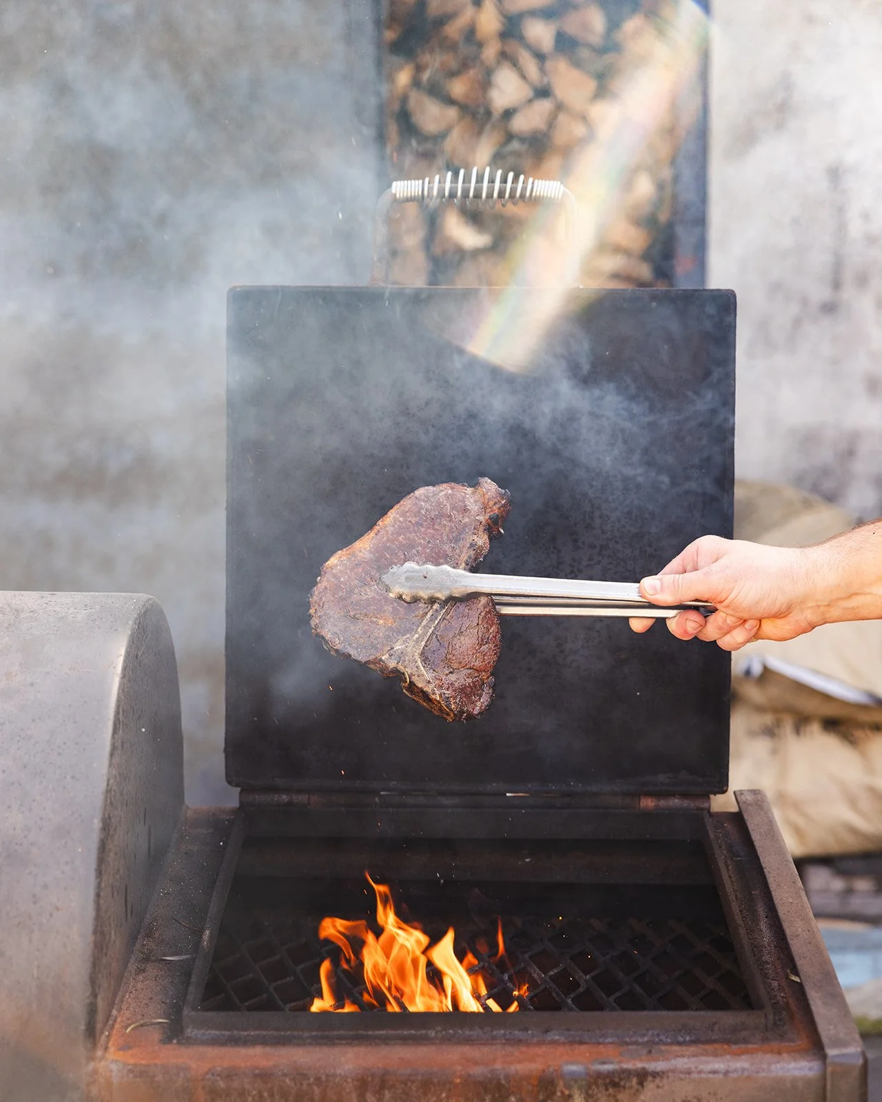 Person using tongs to cook a large piece of meat on an outdoor grill over an open flame.