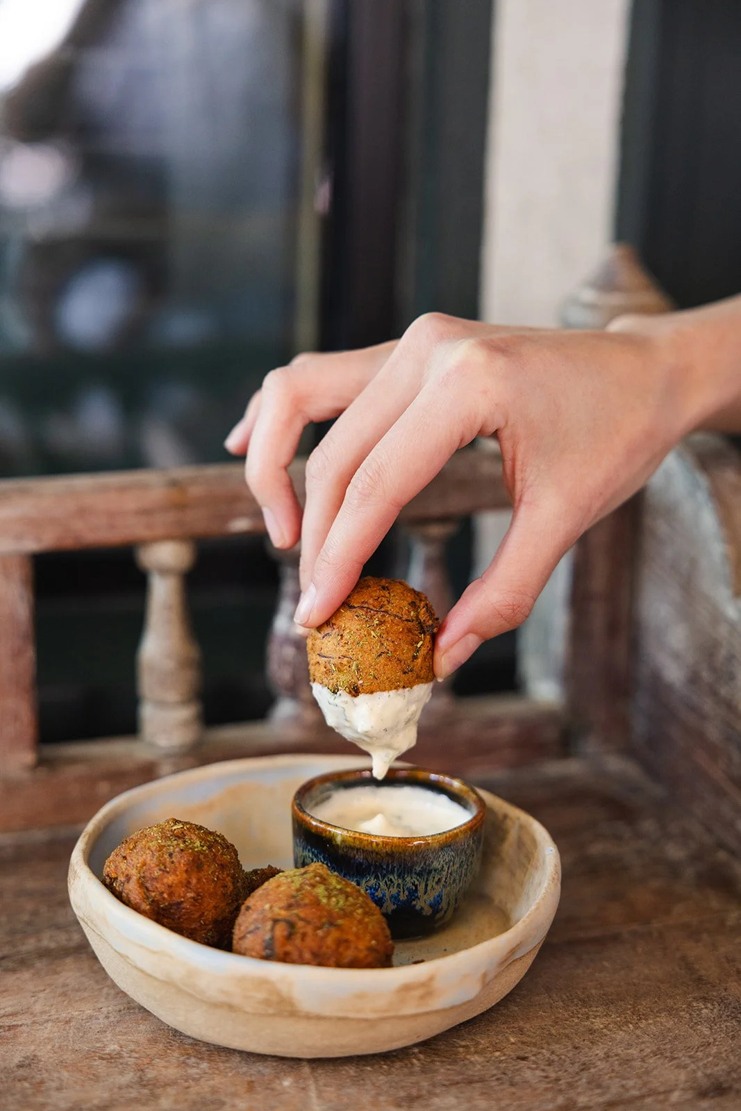 Hand dipping a falafel ball into a small bowl of creamy white sauce on a rustic wooden table, with additional falafel balls in a beige bowl nearby.