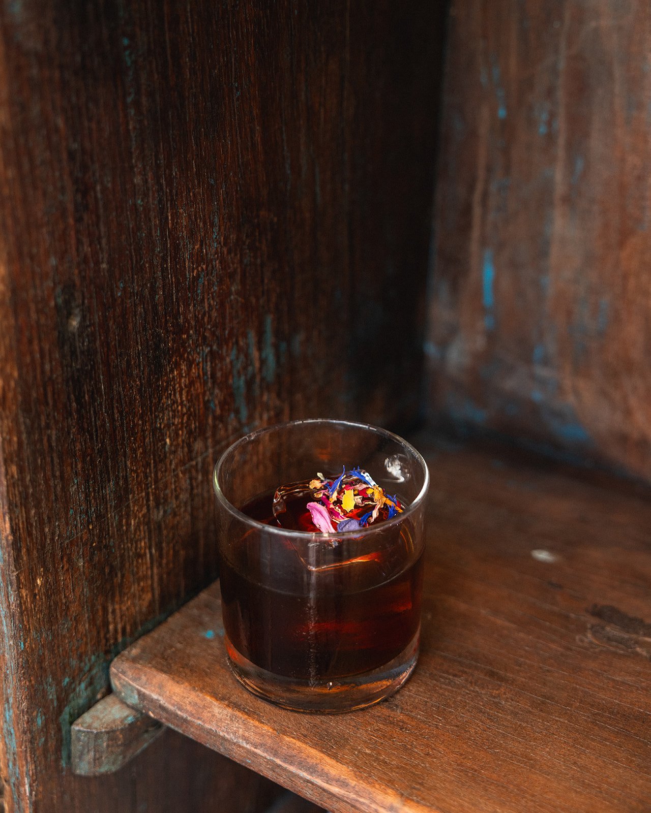 A glass of dark red herbal tea with colorful flower petals floating on top, placed on a wooden shelf inside a rustic wooden cabinet.