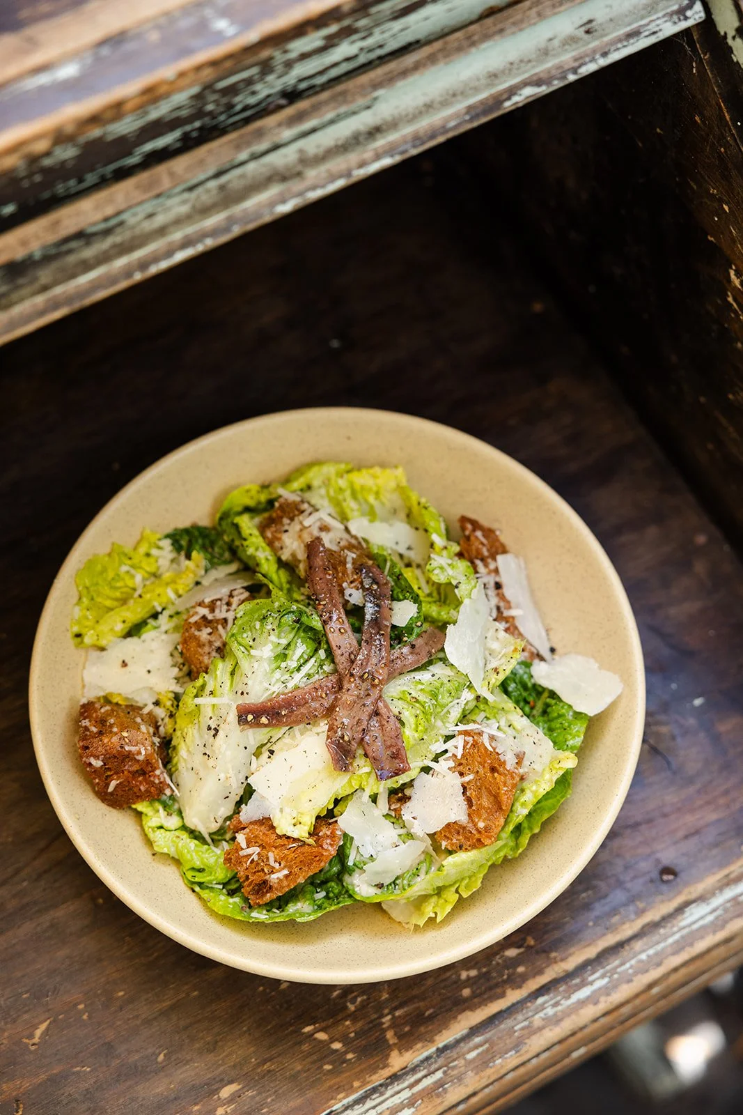 Caesar salad with lettuce, croutons, shredded cheese, anchovies, and grated cheese in a beige bowl on a wooden table.