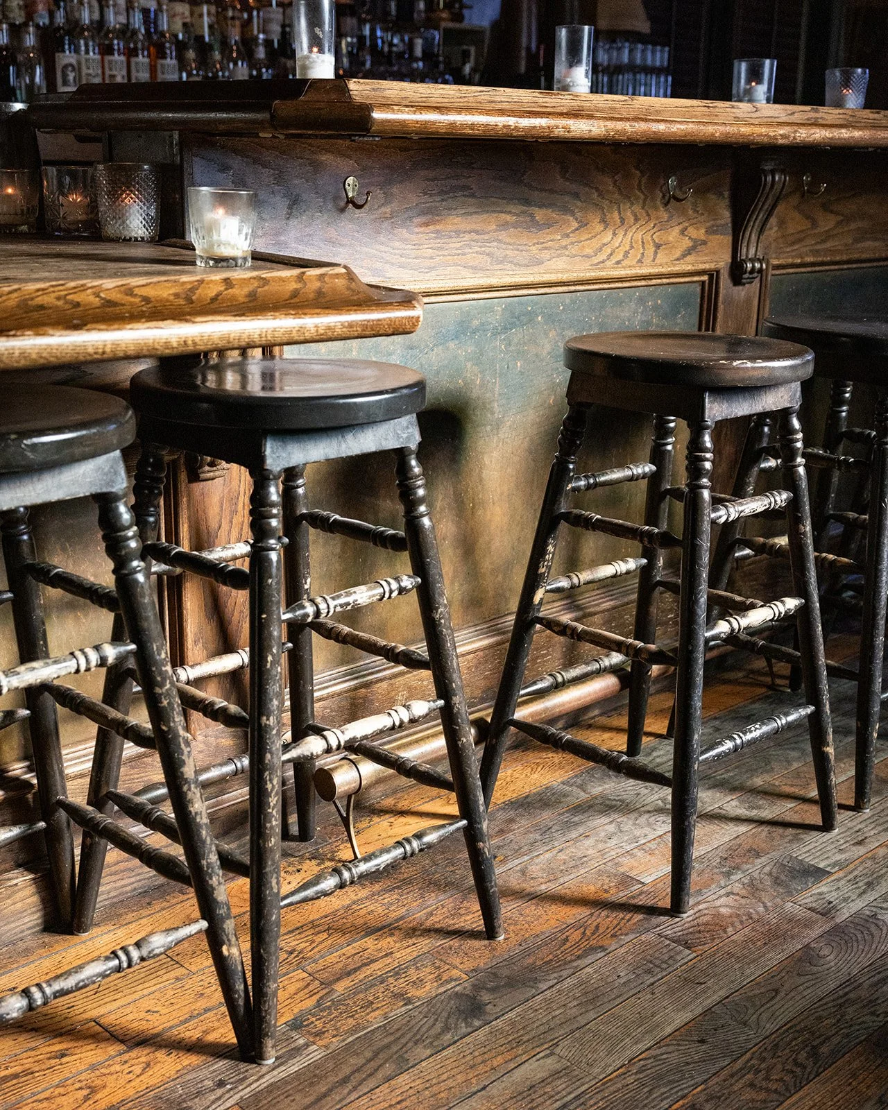 Three black wooden bar stools with worn paint, placed in front of a rustic wooden bar counter in a dimly lit bar or restaurant.