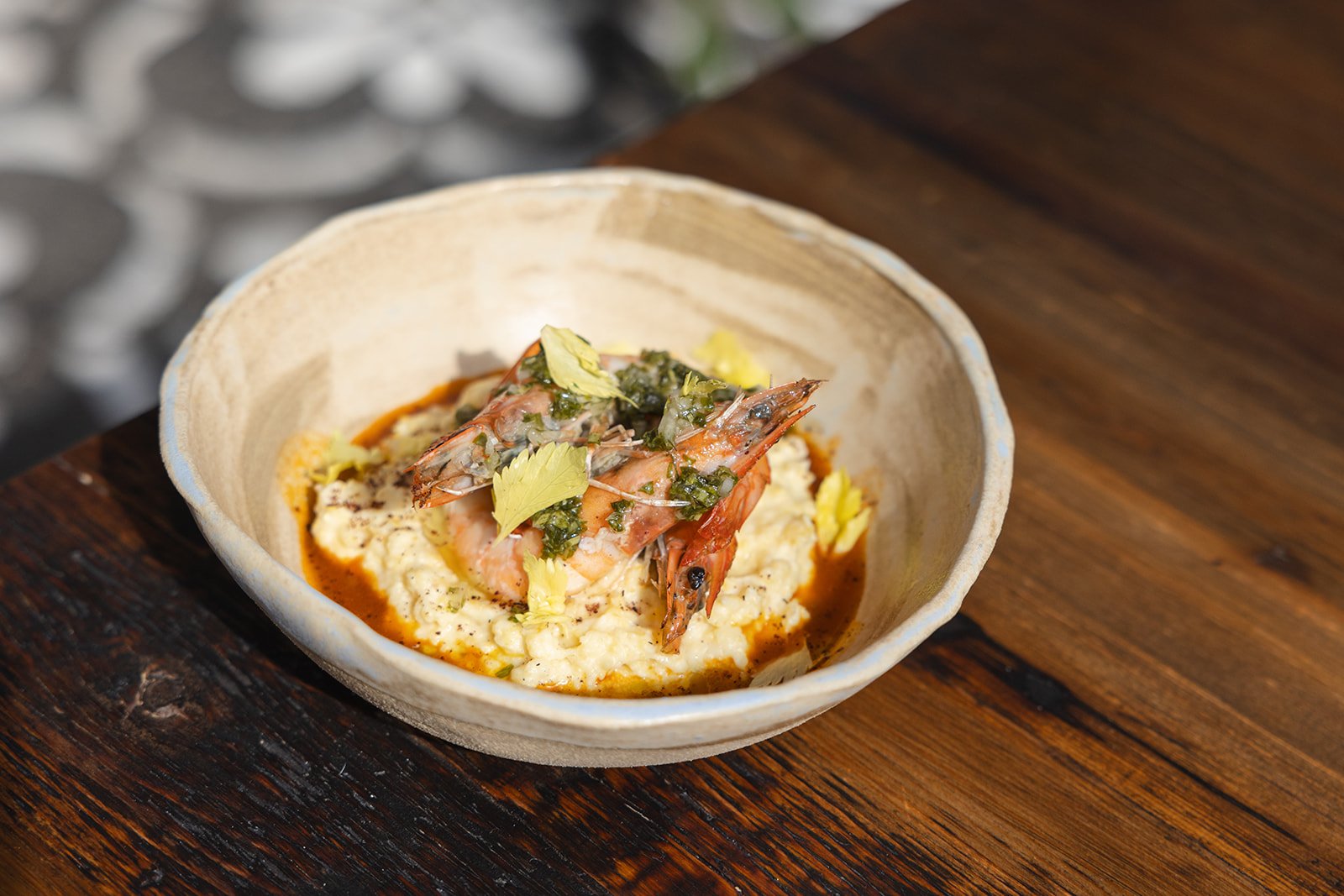 A bowl of shrimp and sauce served over a creamy grain or vegetable puree, garnished with herbs and green oil.