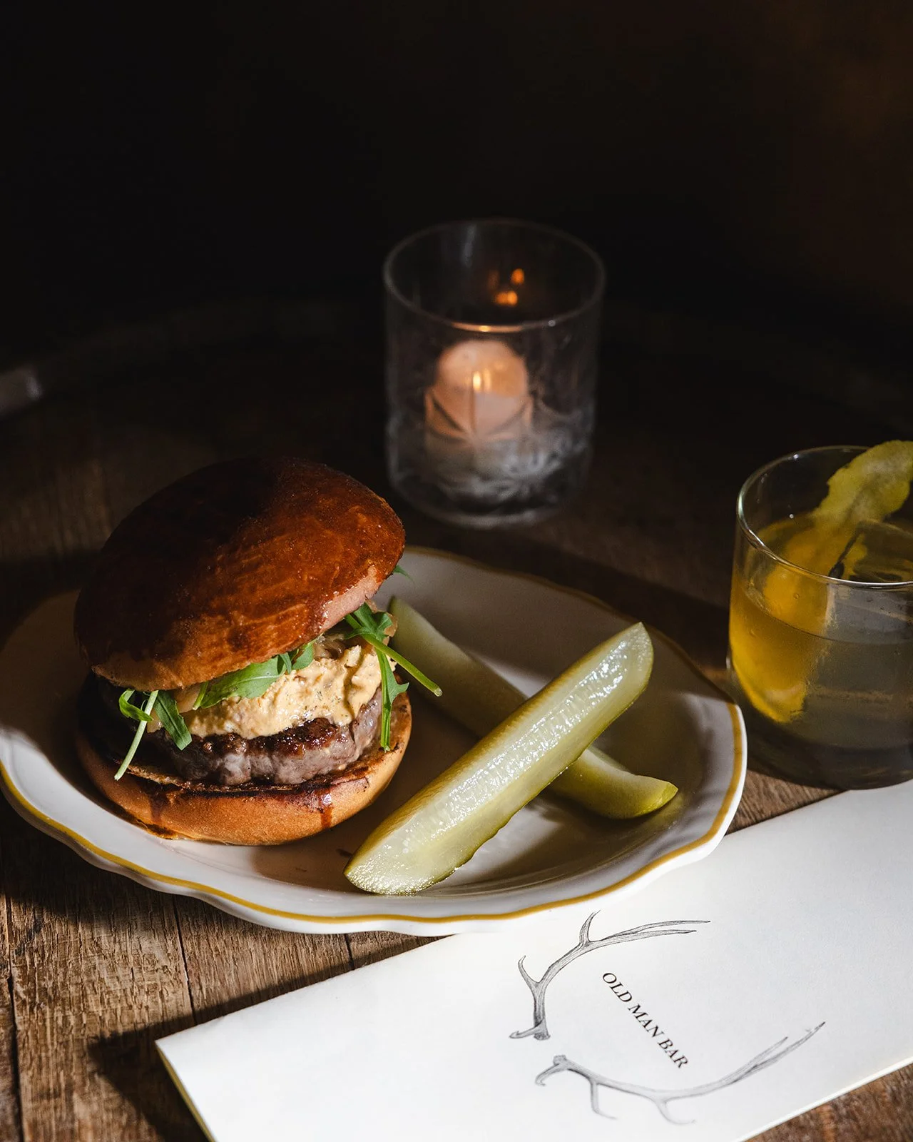 A burger with greens and sauce on a plate, served with pickle spears, a lit candle, a glass of yellow drink with ice, and a menu that reads 'Old Man Bar' on a wooden table.