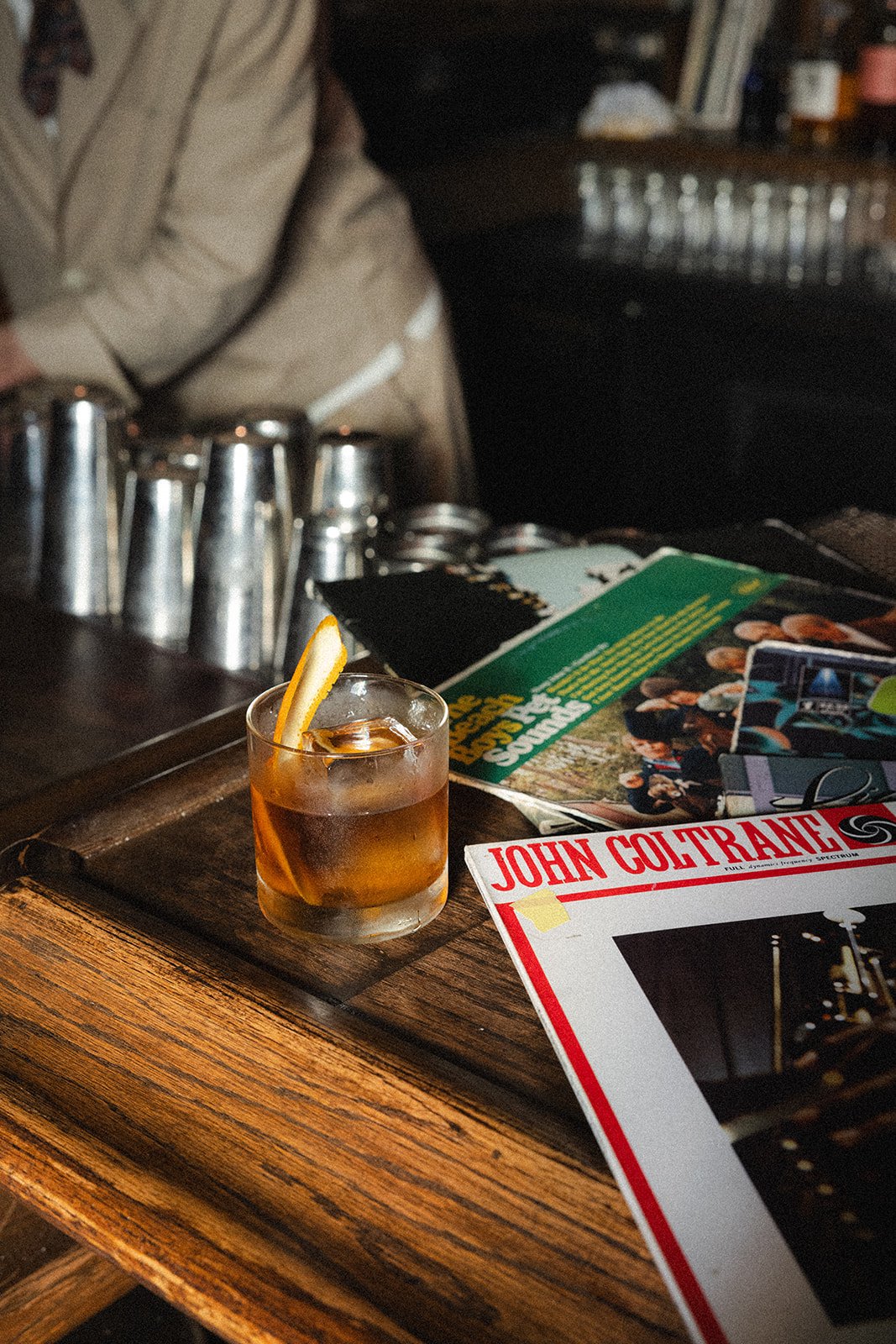 A glass of whiskey with an ice cube and lemon peel garnish on a wooden table, surrounded by magazines and a person in a beige jacket.