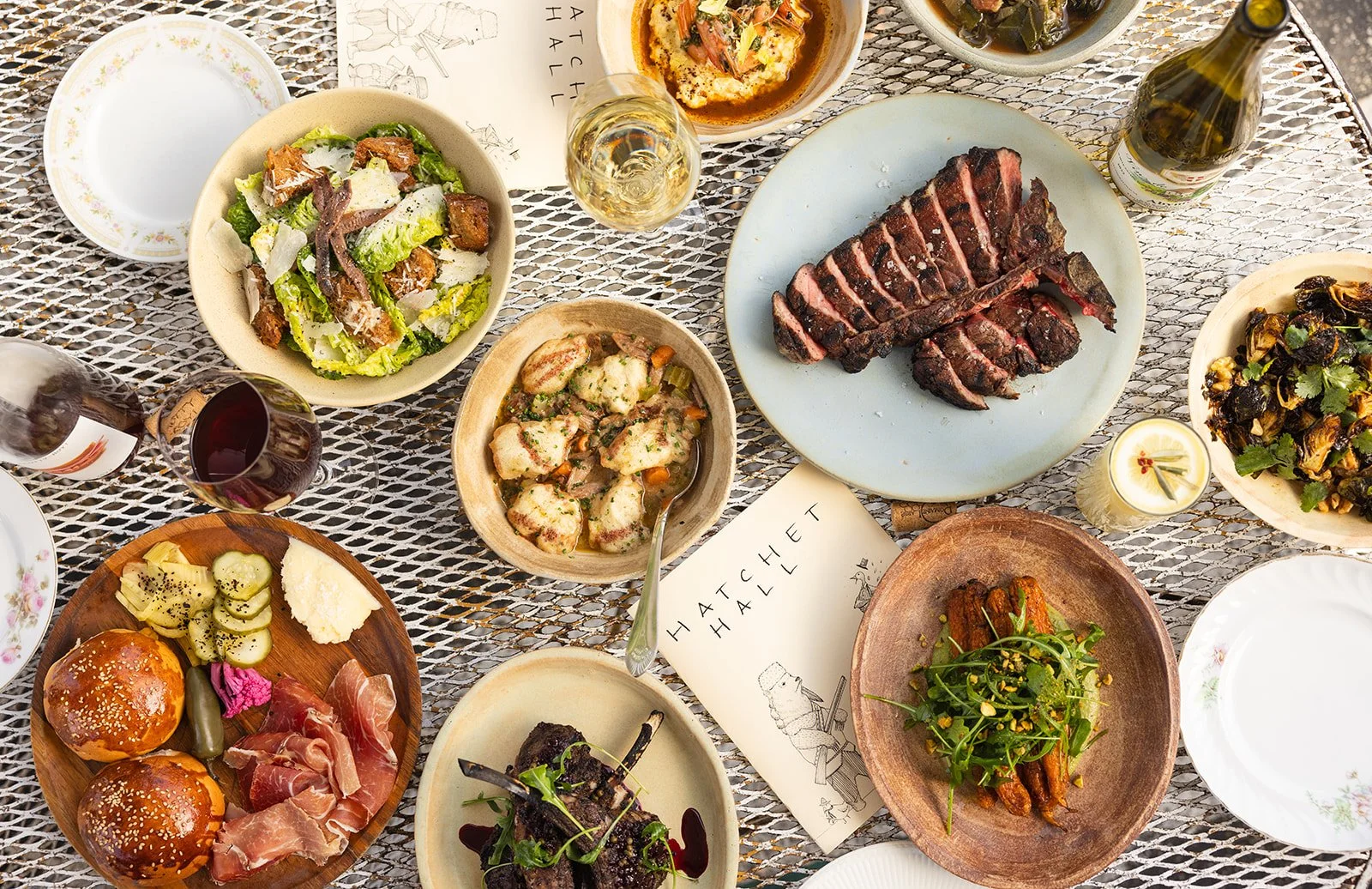 A diverse spread of dishes on a table including salad, bread rolls with accompaniments, grilled steak, roasted vegetables, fish with sauce, and various salads, along with glasses of wine and a small candle.