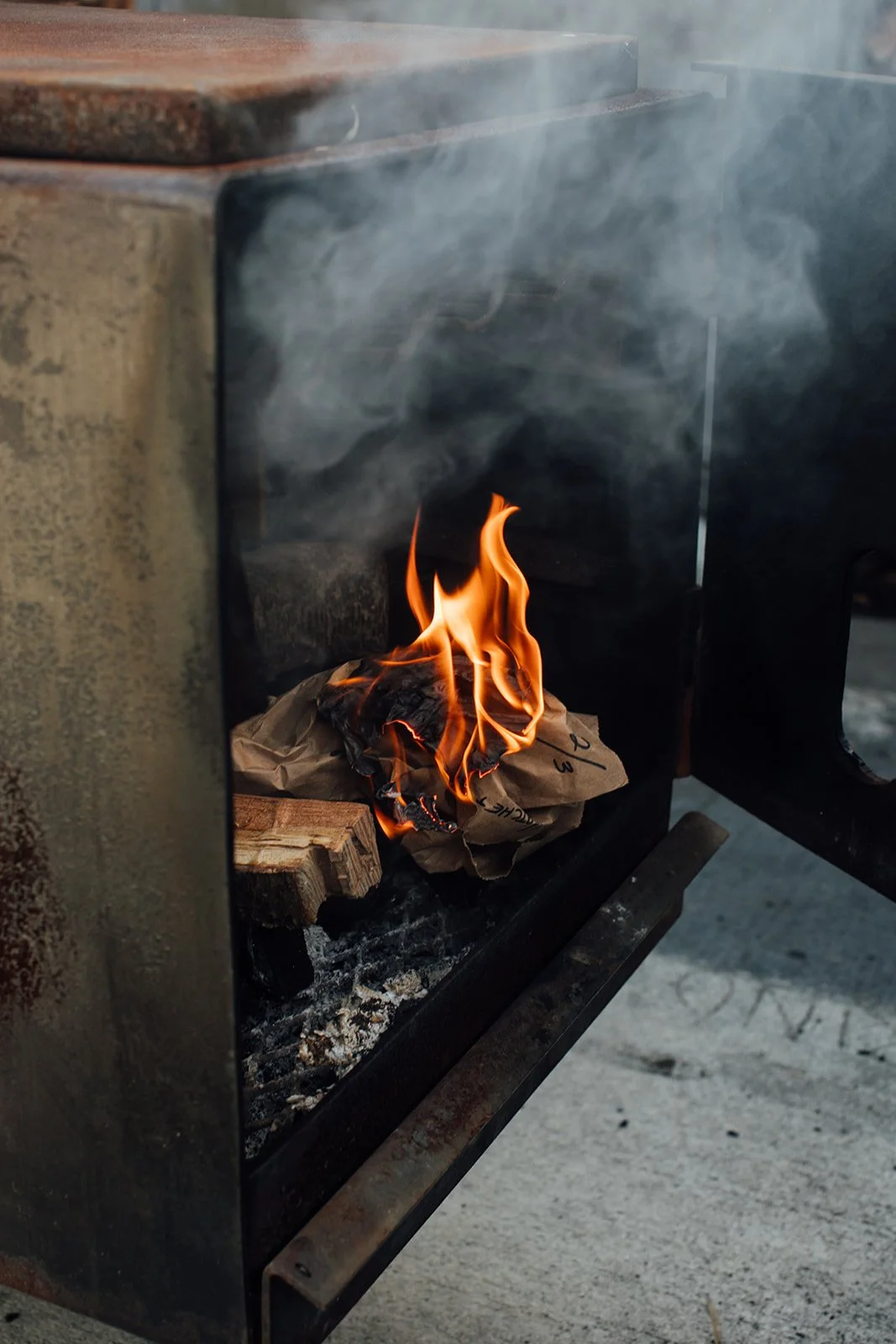 Close-up of a small fire burning inside a metal stove, with flames rising over crumpled paper and wood pieces.