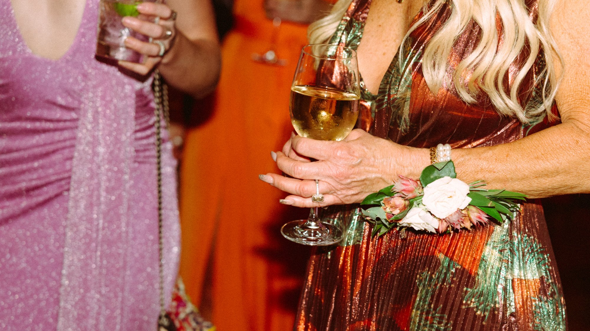 Close-up of a woman holding a glass of white wine at a party, wearing a floral dress with a white flower corsage and a pearl bracelet. Another woman in a pink sparkly dress is partially visible in the background.