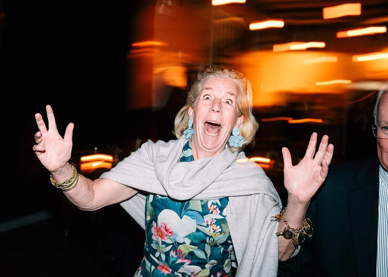 A woman with blonde hair, wearing blue earrings, a floral dress, and gold bracelets, appears surprised or excited with her mouth open and hands raised, at a lively event with warm lighting in the background.
