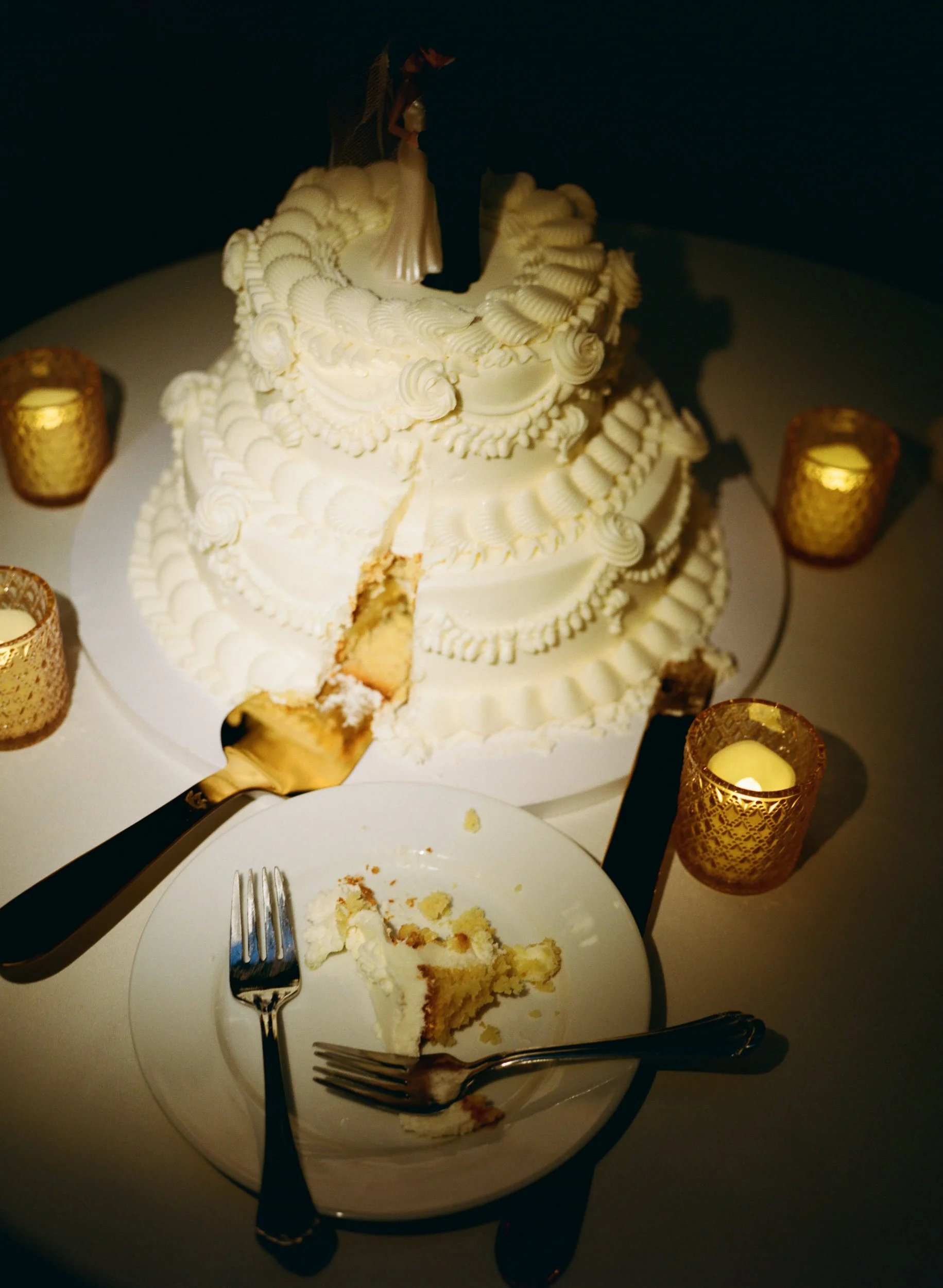 Round, multi-tiered wedding cake with white icing and ornate piping, a bride and groom figurine on top, with a slice removed showing the interior, on a table with four gold candle holders.