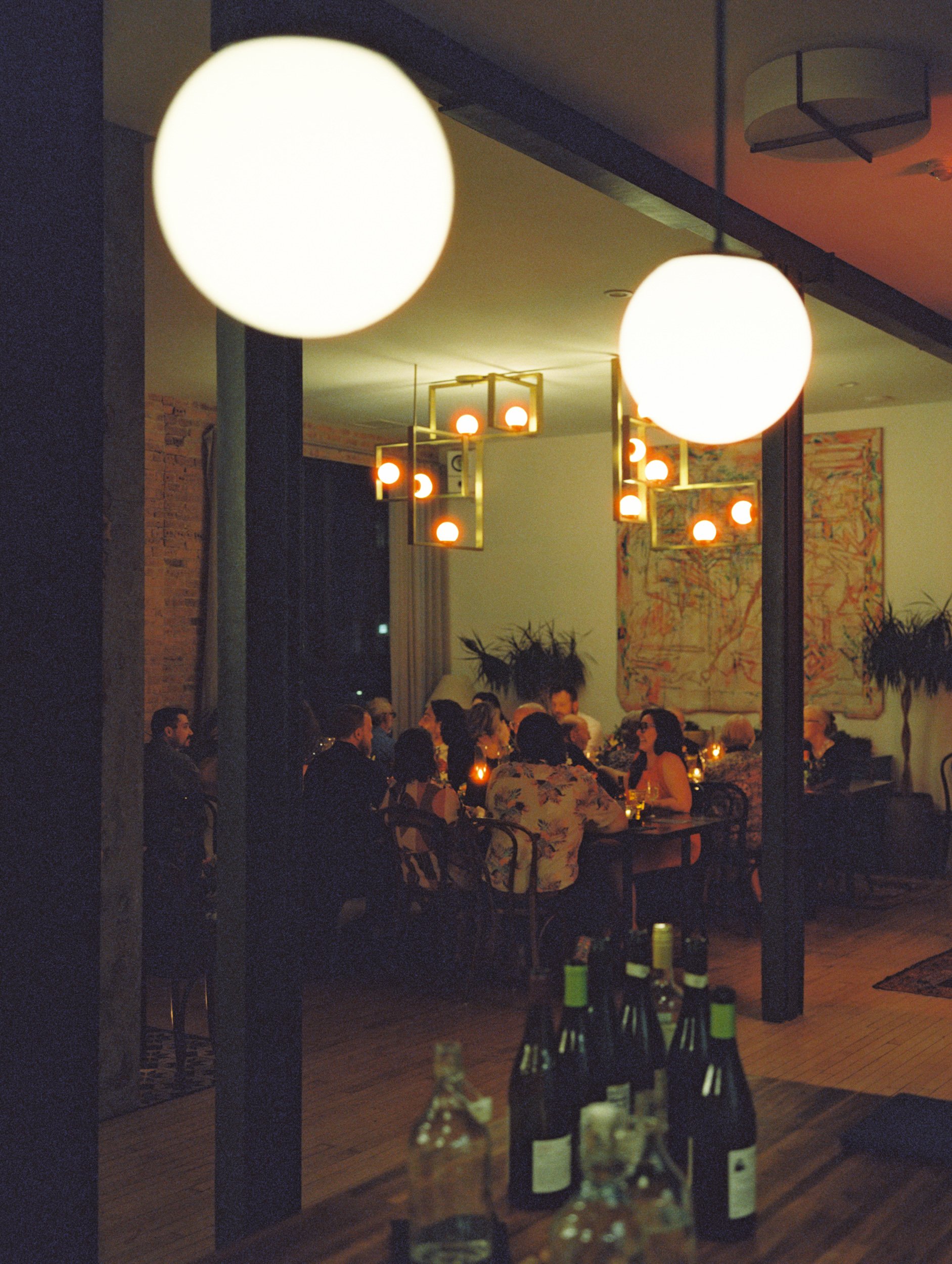 People dining at a restaurant during the evening, illuminated by hanging globe and modern pendant lights, with bottles on the table in the foreground.