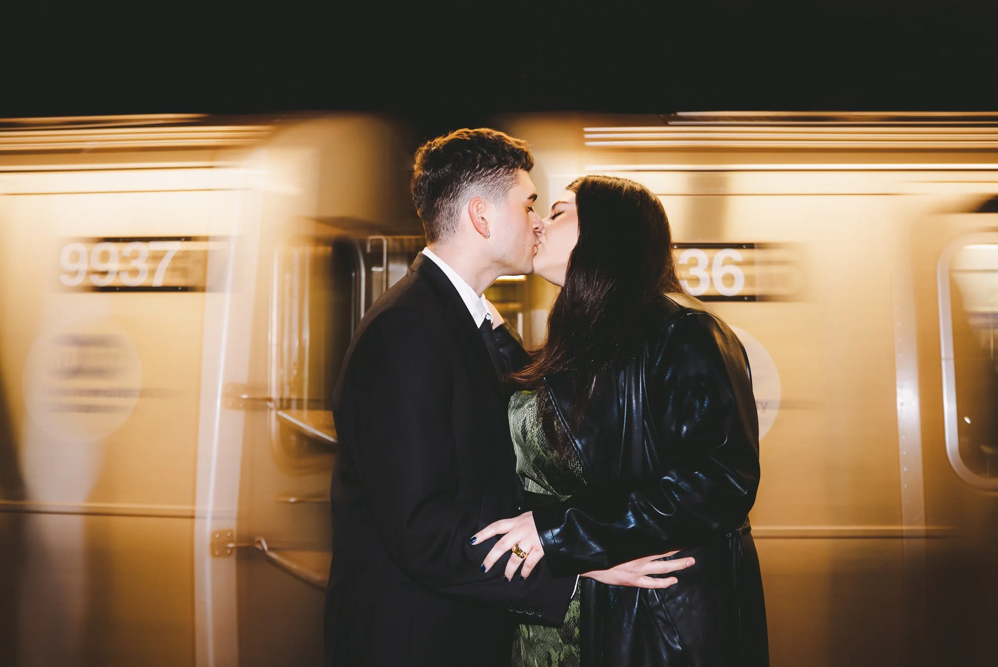 A young couple in formal attire sharing a kiss on a subway platform at night, with a moving subway train in the background.