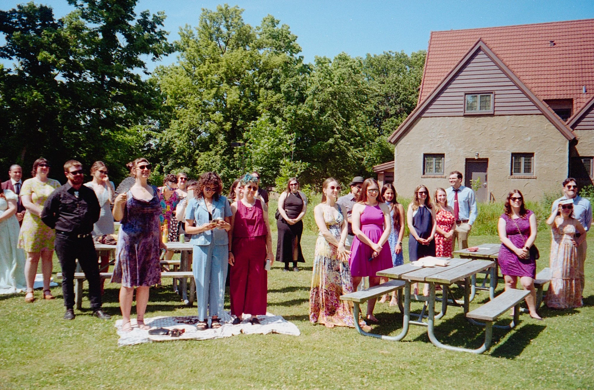 A group of people gathered outdoors on a grassy lawn during a sunny day, with some standing on a blanket and others near a picnic table, in front of a house with a red roof and trees in the background.