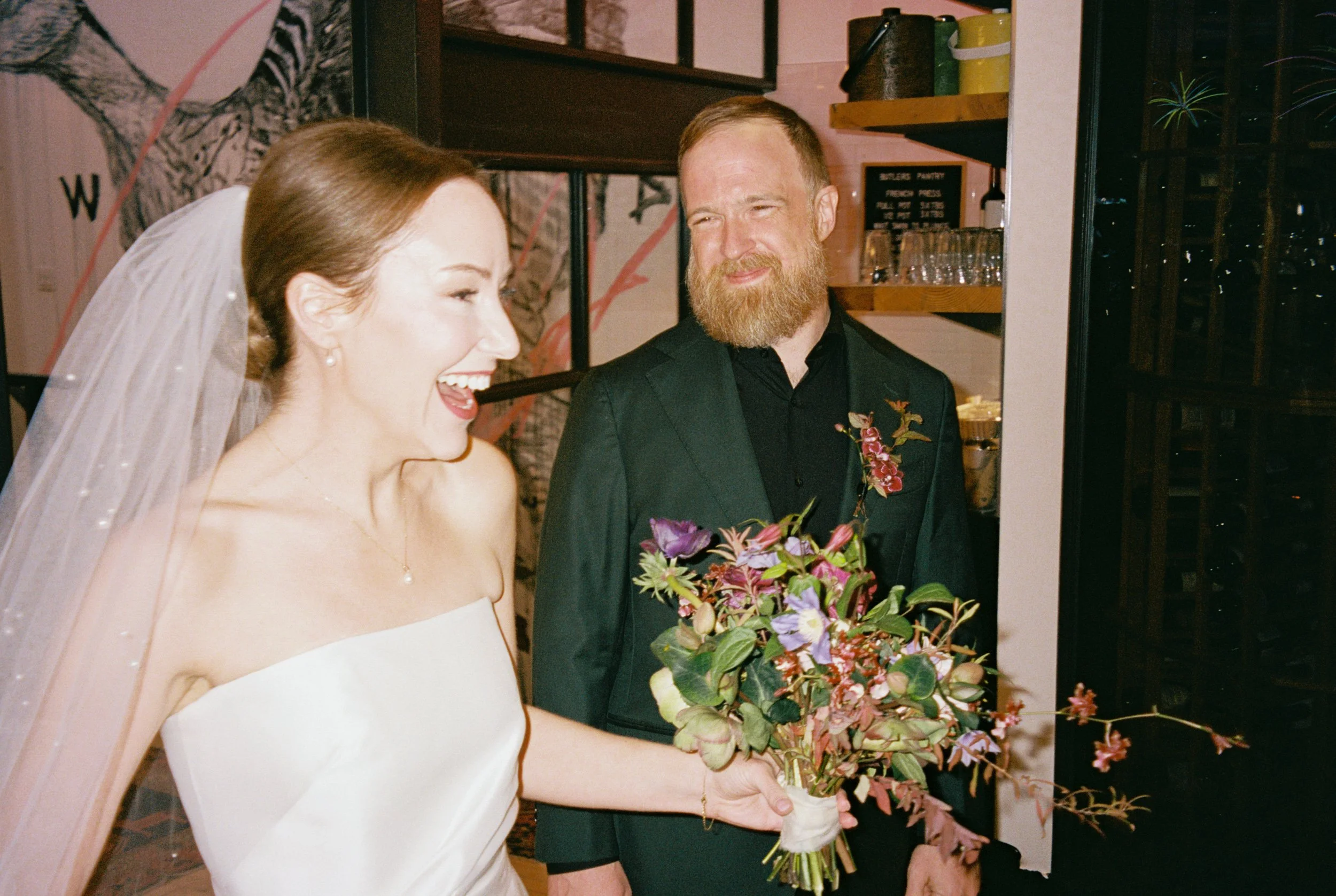 A bride with a veil and strapless dress holding a bouquet of flowers and smiling, standing next to a bearded man in a dark suit, inside a decorated venue.