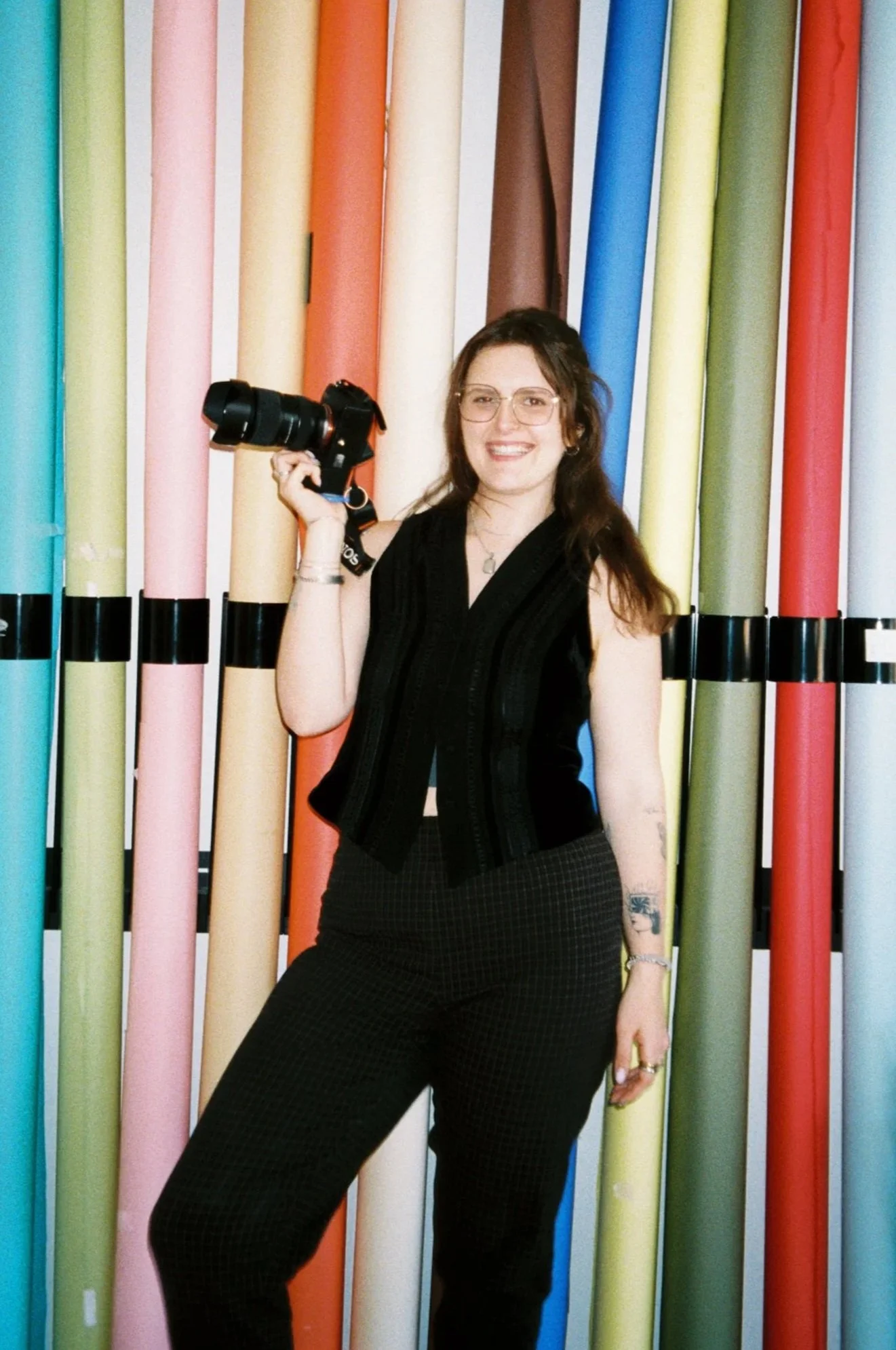 A woman smiling and holding a camera stands in front of a colorful background of large rolled papers in pastel and bright colors.