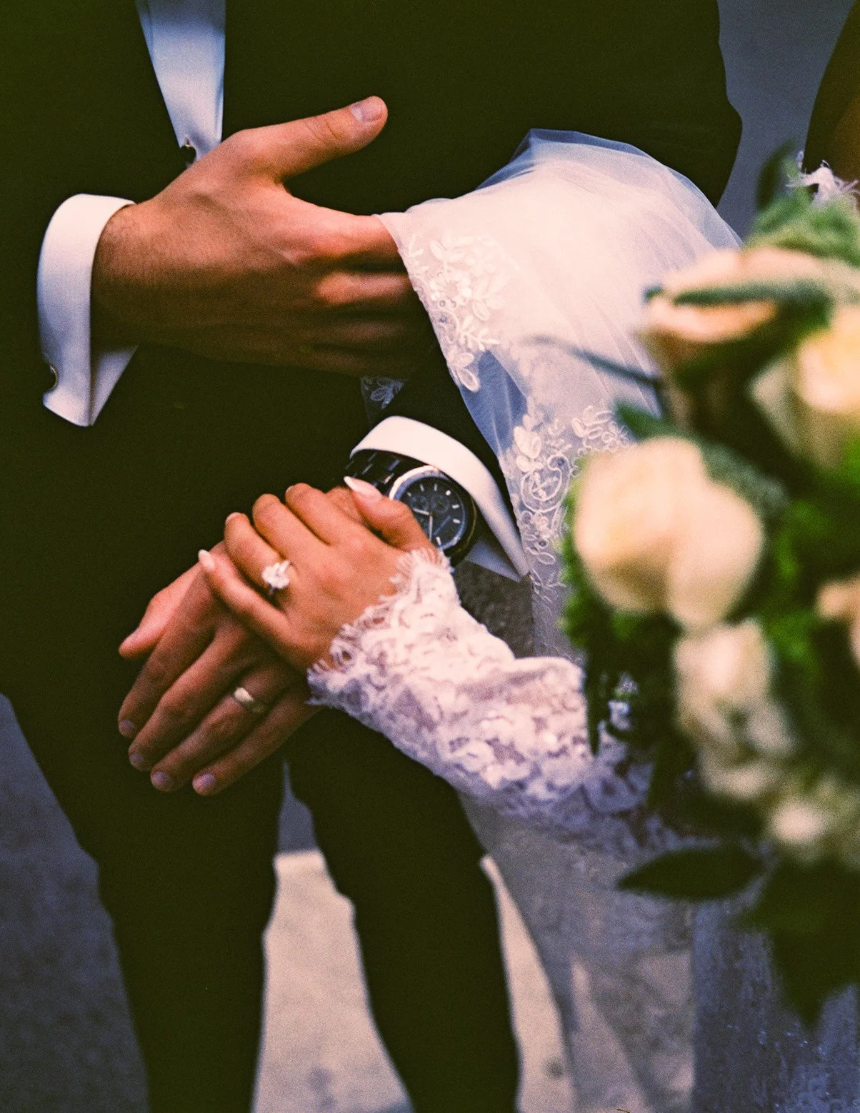 Close-up of a bride and groom holding hands, showing wedding rings. The bride's hand rests on the groom's arm, which is wearing a watch, and the groom's hand is gently holding the bride's hand. The groom is wearing a watch and a suit with a bow tie, 