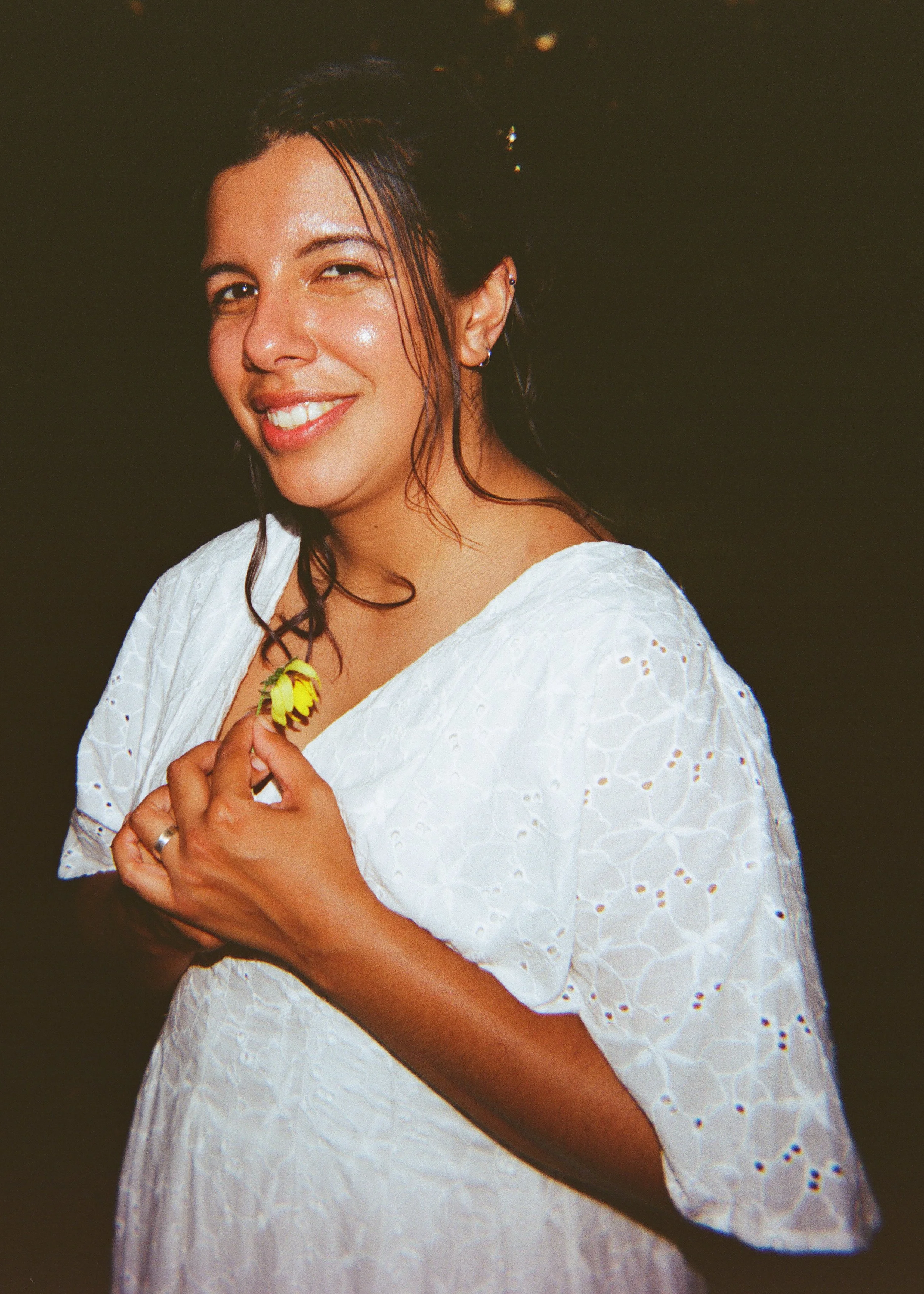 A woman smiling and winking, wearing a white dress with eyelet details, holding a yellow flower near her chest.
