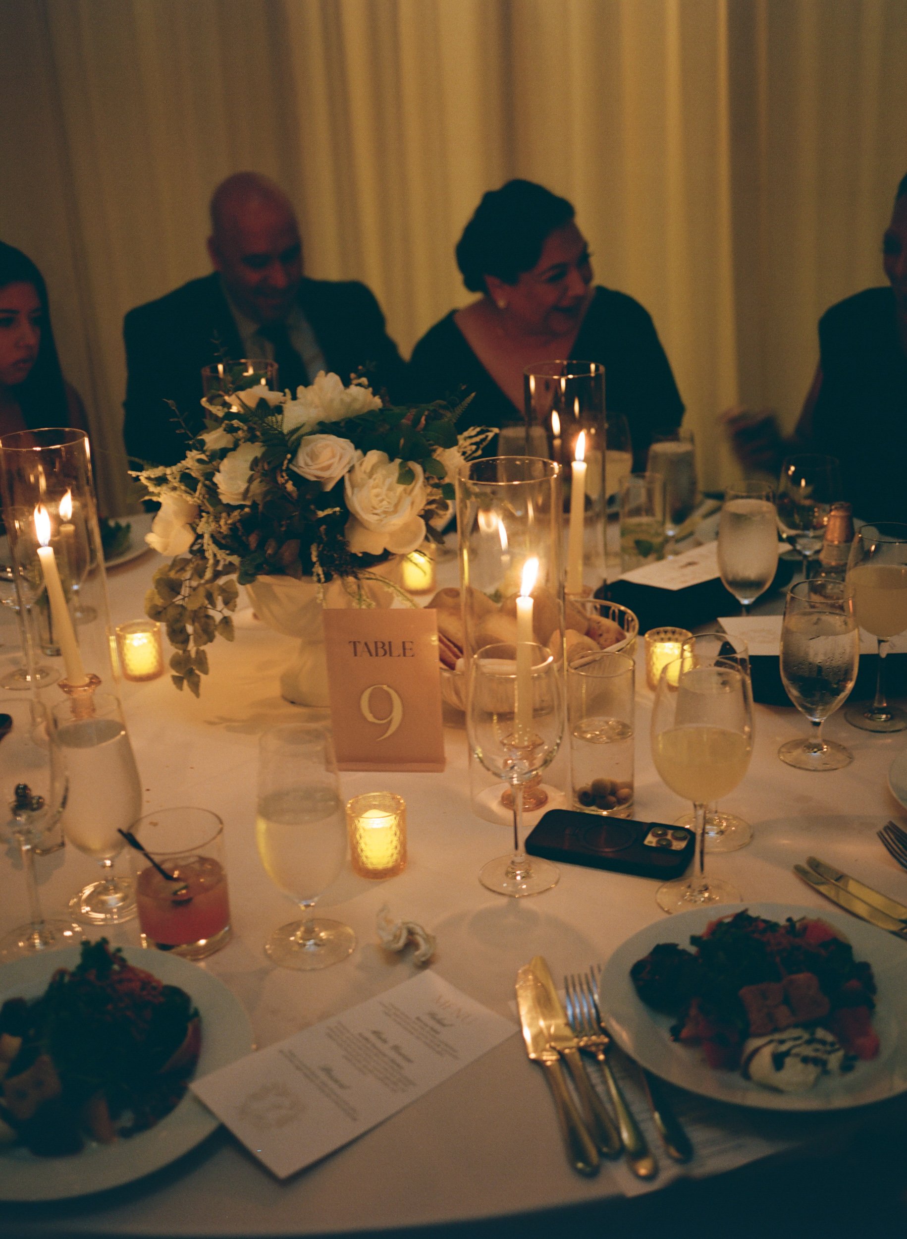 A formal dinner table with a floral centerpiece, candles, glasses of white wine, plates with food, and a menu. People are seated in the background. Table number 9 is visible.