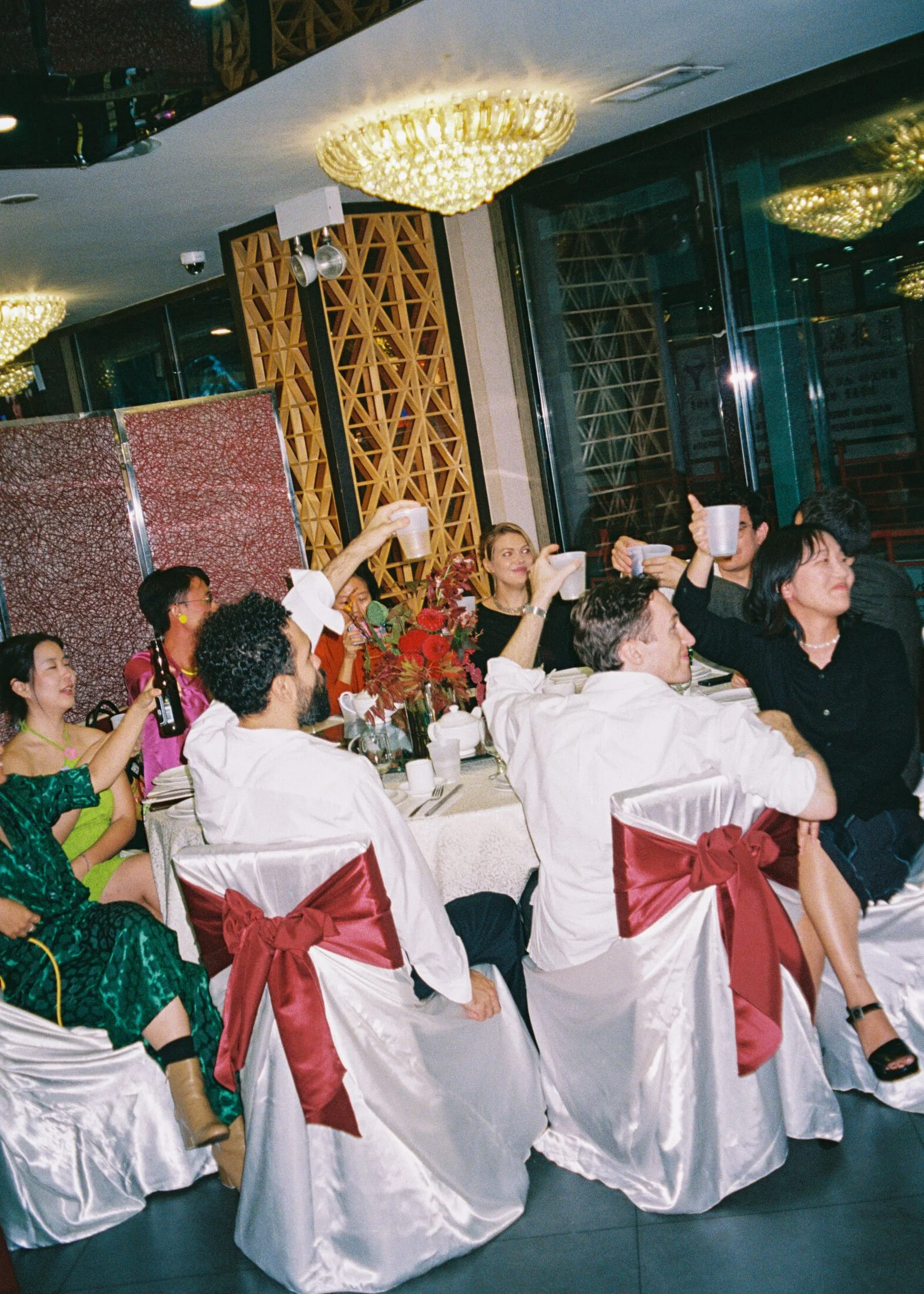 People celebrating at a banquet table with drinks, a floral centerpiece, white chairs with red bows, and a decorated background with chandeliers and a large window.