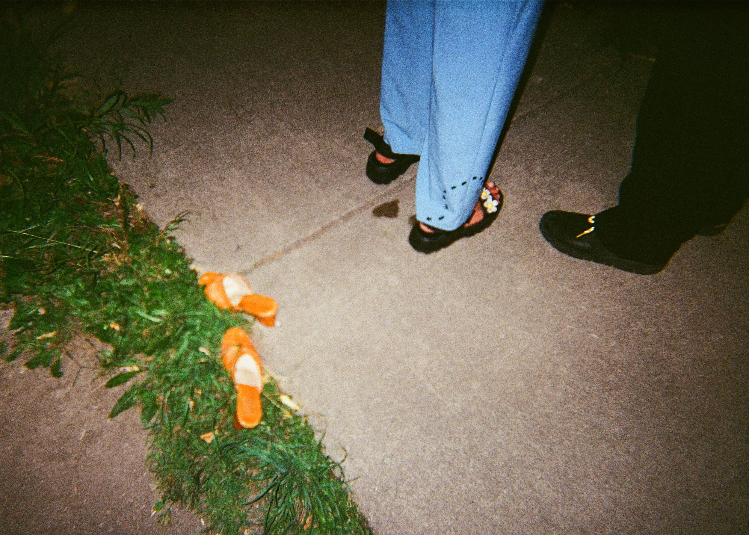 Close-up of the lower half of two people standing on a sidewalk, with orange and white mushroom decorations on a green plant nearby.