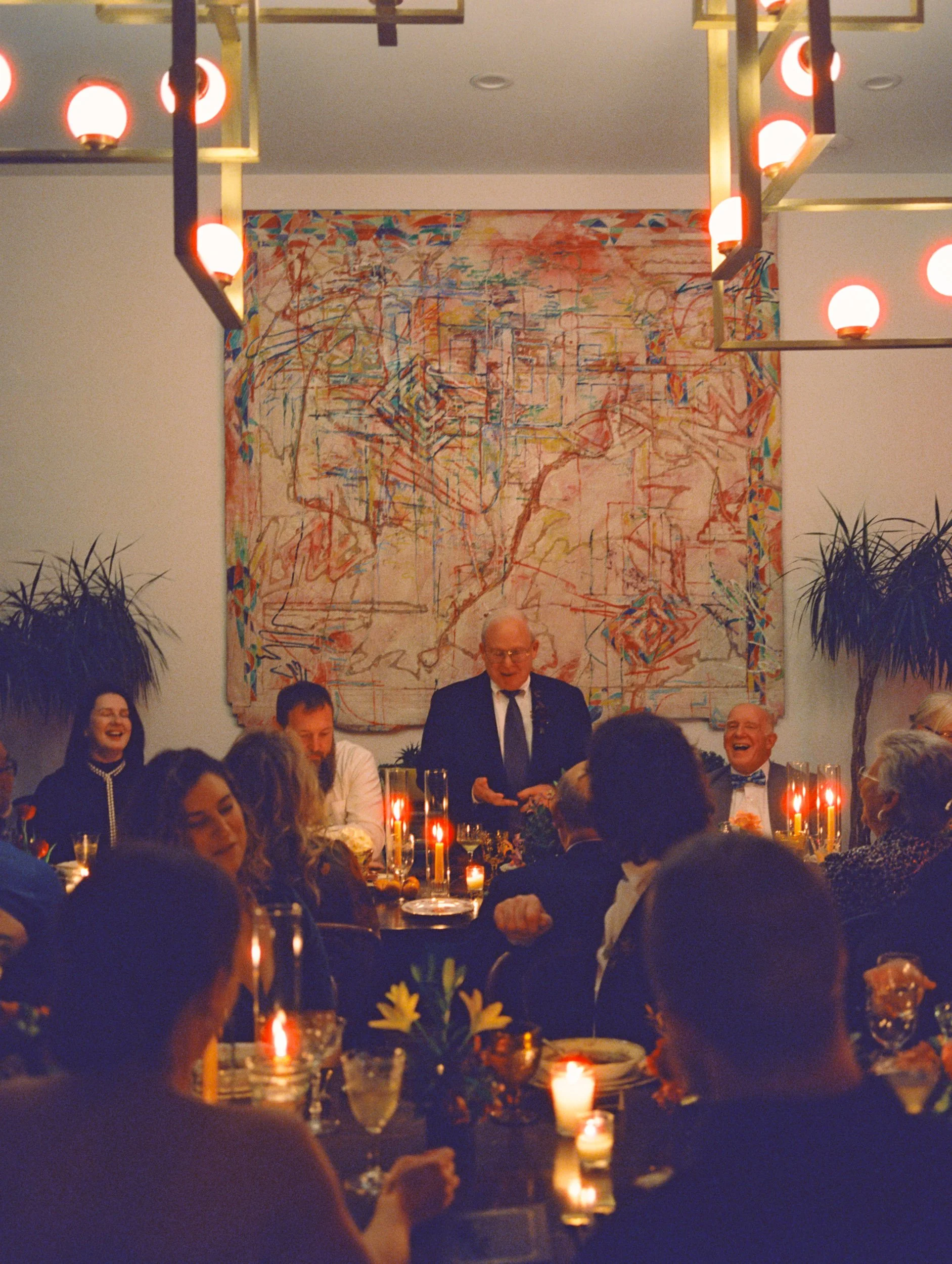 A group of people gathered around a dinner table in a warmly lit room with candles, with an elderly man in a suit speaking or giving a toast. A large abstract painting hangs on the wall behind him, and plants decorate the room.