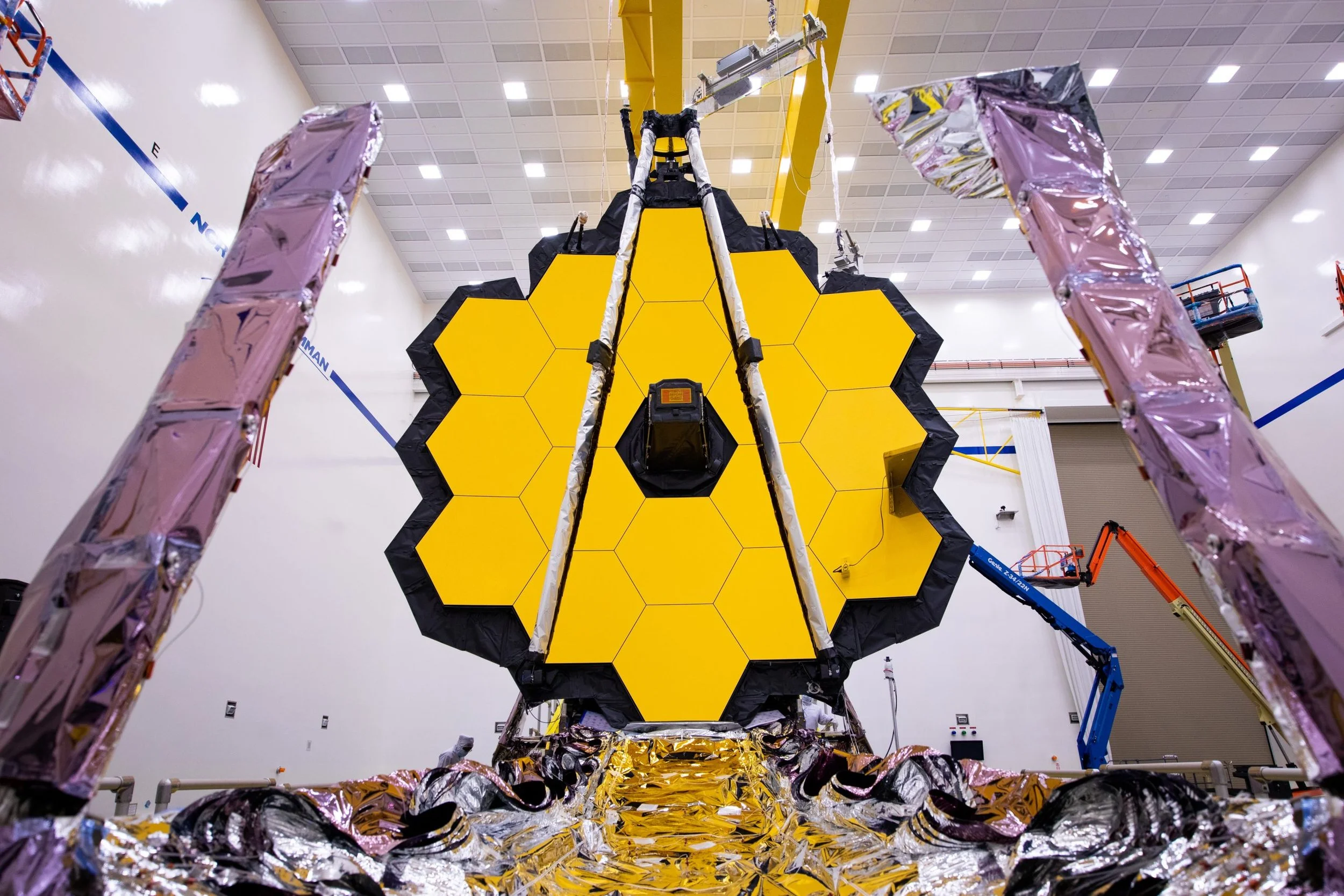 Large spacecraft with a yellow and black honeycomb-patterned surface inside a spacious industrial facility, surrounded by scaffolding and maintenance equipment.