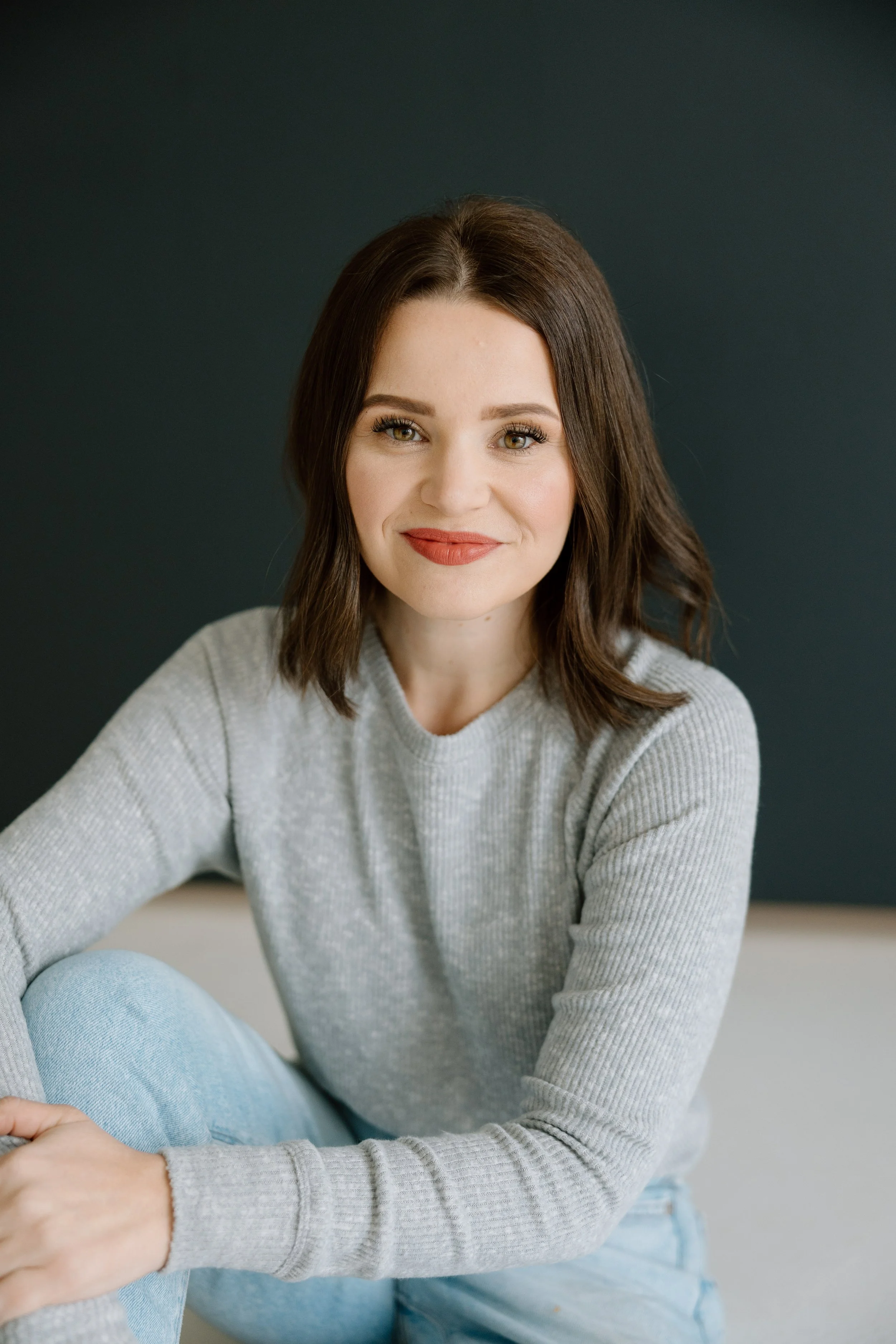 A woman with shoulder-length brown hair, wearing a light gray sweater, sitting against a dark gray background, smiling at the camera.