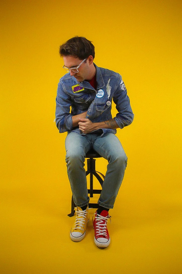 A young man wearing glasses, a denim jacket with colorful patches, and light blue jeans, sitting on a black stool against a solid yellow background. He is wearing mismatched sneakers: one yellow and one red, and looking down with his hands clasped over his stomach.