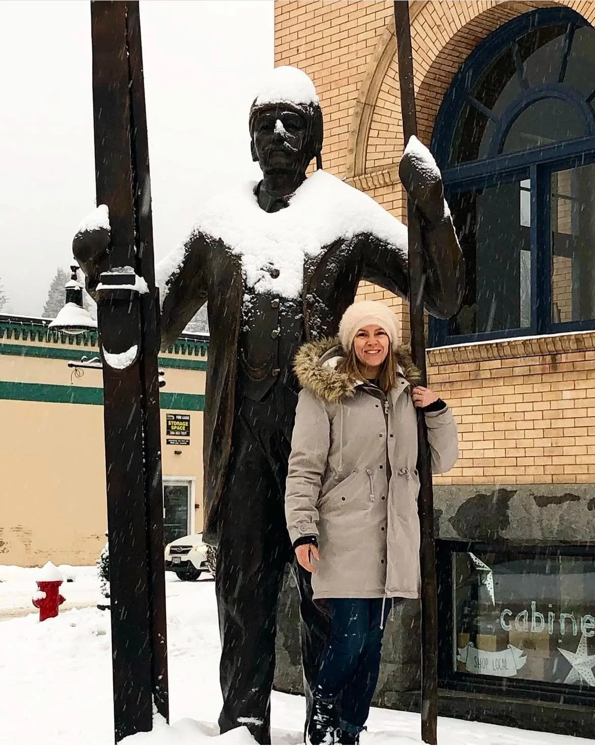 A woman standing outdoors in snowy weather, smiling and dressed in a light-colored winter coat with a fur-lined hood, next to a large statue of a soldier holding a flag, with snow on the statue's shoulders and helmet.