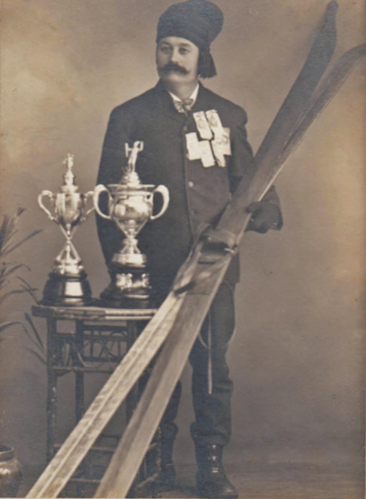 A man in historical attire with a beard and mustache, wearing a turban, standing next to a table with three trophies, holding a large wooden plank or beam.