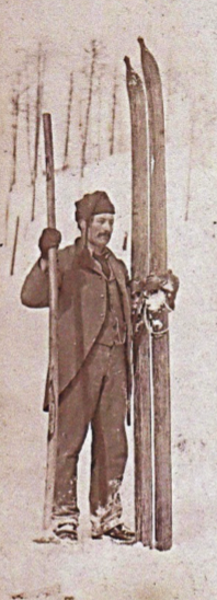 A man in vintage clothing standing outdoors, holding a pair of skis and a ski pole.