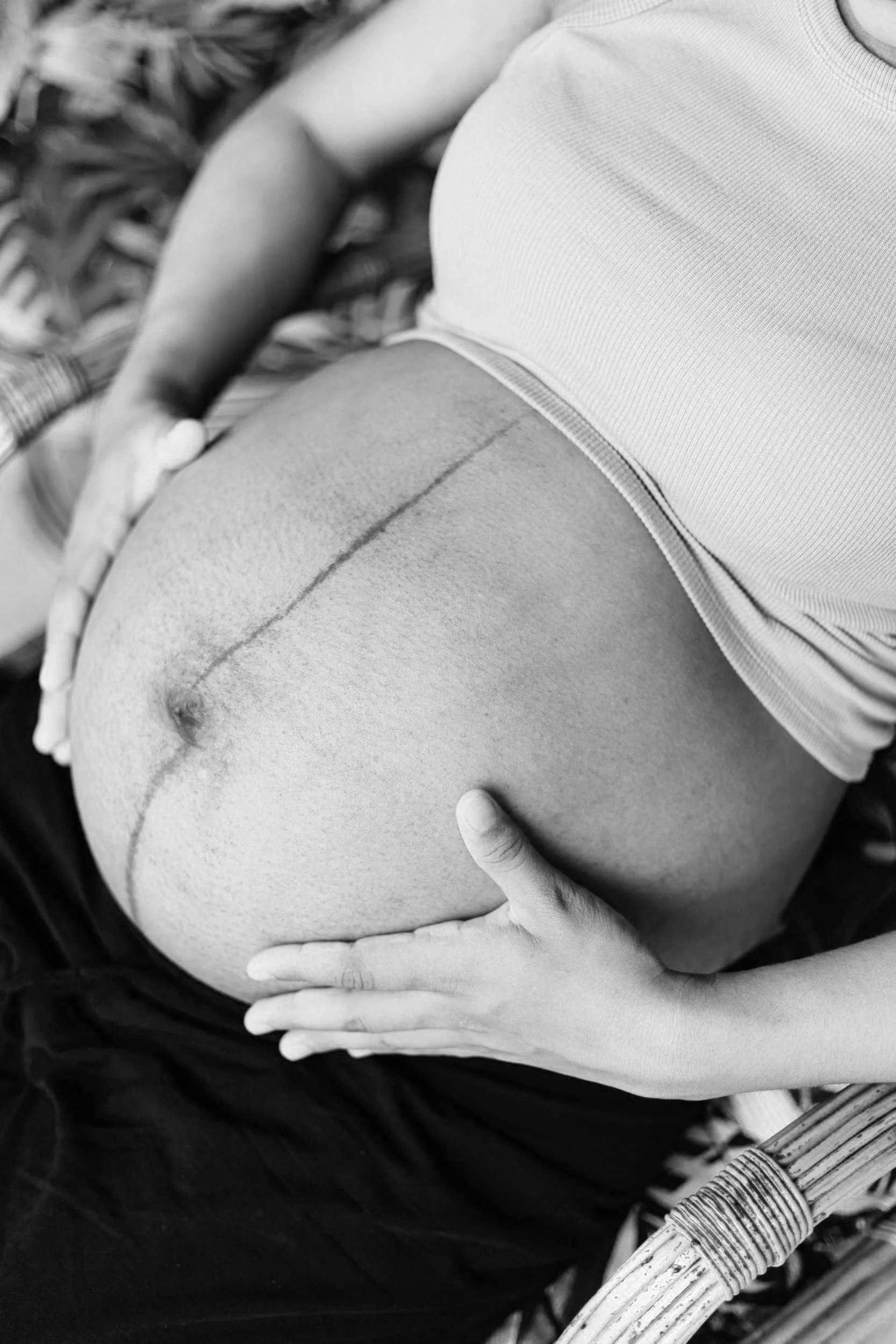Close-up of a pregnant woman's bare belly with a line drawn on it, hands resting on the belly, wearing a light-colored shirt, outdoors with plants or foliage in the background.