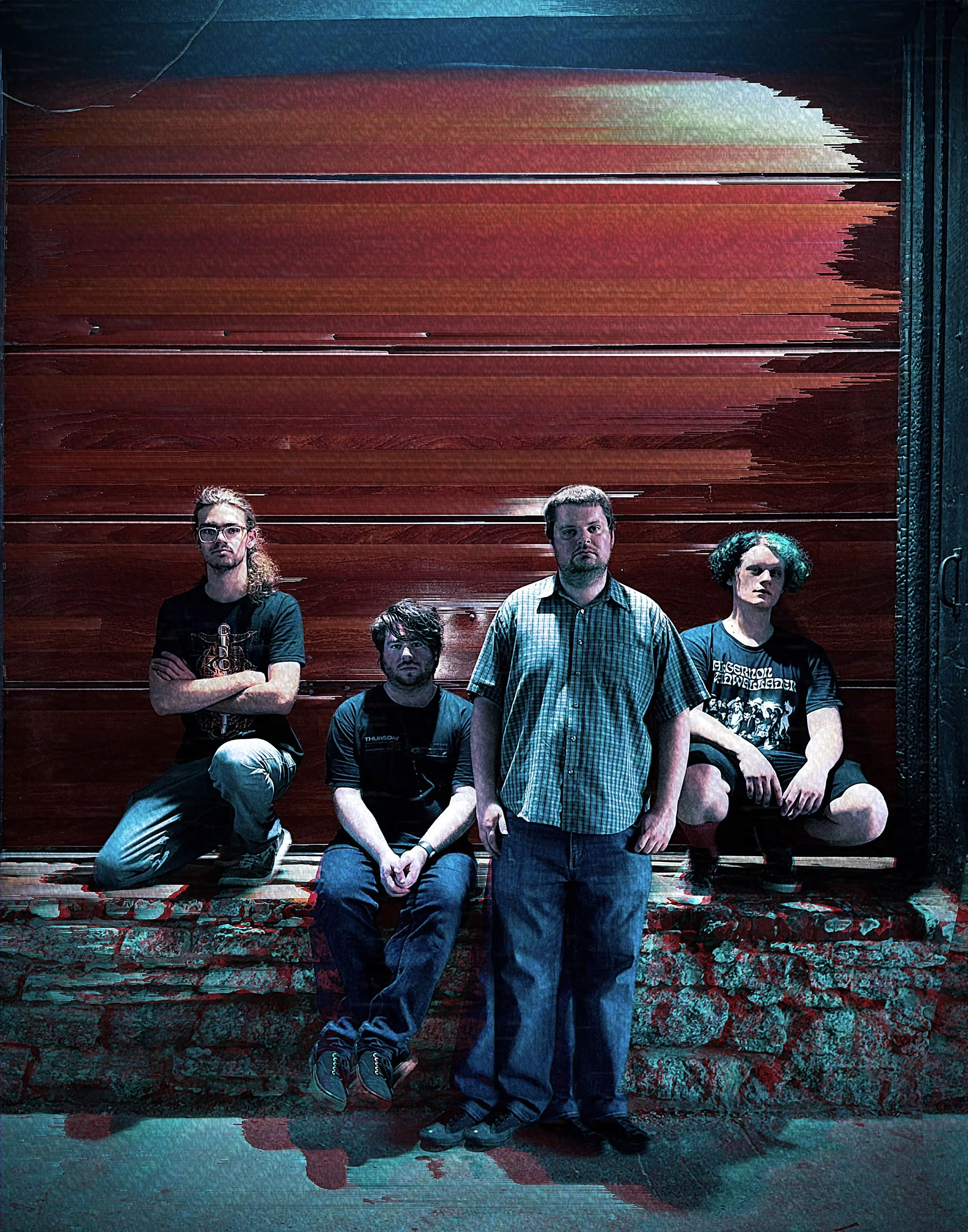 A group of five individuals, four men and one woman, standing and sitting in front of a large wooden garage door with visible damage. The man in the center is standing with his hands at his sides, wearing a checkered shirt. The other four people have serious expressions; one man has long hair and glasses, another is sitting with crossed arms, another has short hair and is standing behind the man in the checkered shirt, and the woman has curly hair and is sitting with her arm resting on her knee. They are positioned on a red brick surface with a worn, textured wall at the bottom of the image.