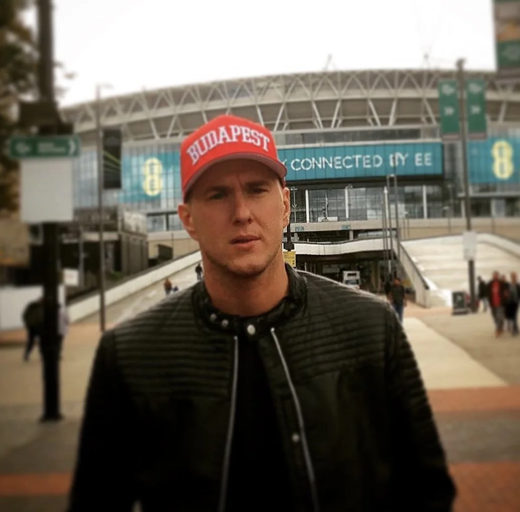 A man wearing a red cap labeled 'BUDAPEST' standing outside a modern stadium or convention center with a digital sign that reads 'CONNECTED IN EE' in the background.