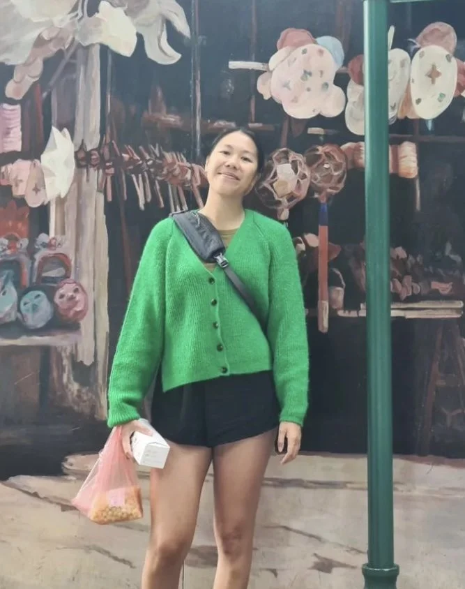 Woman standing in front of a painted market scene, wearing a green cardigan, black shorts, holding snacks and a drink, with a black shoulder bag.