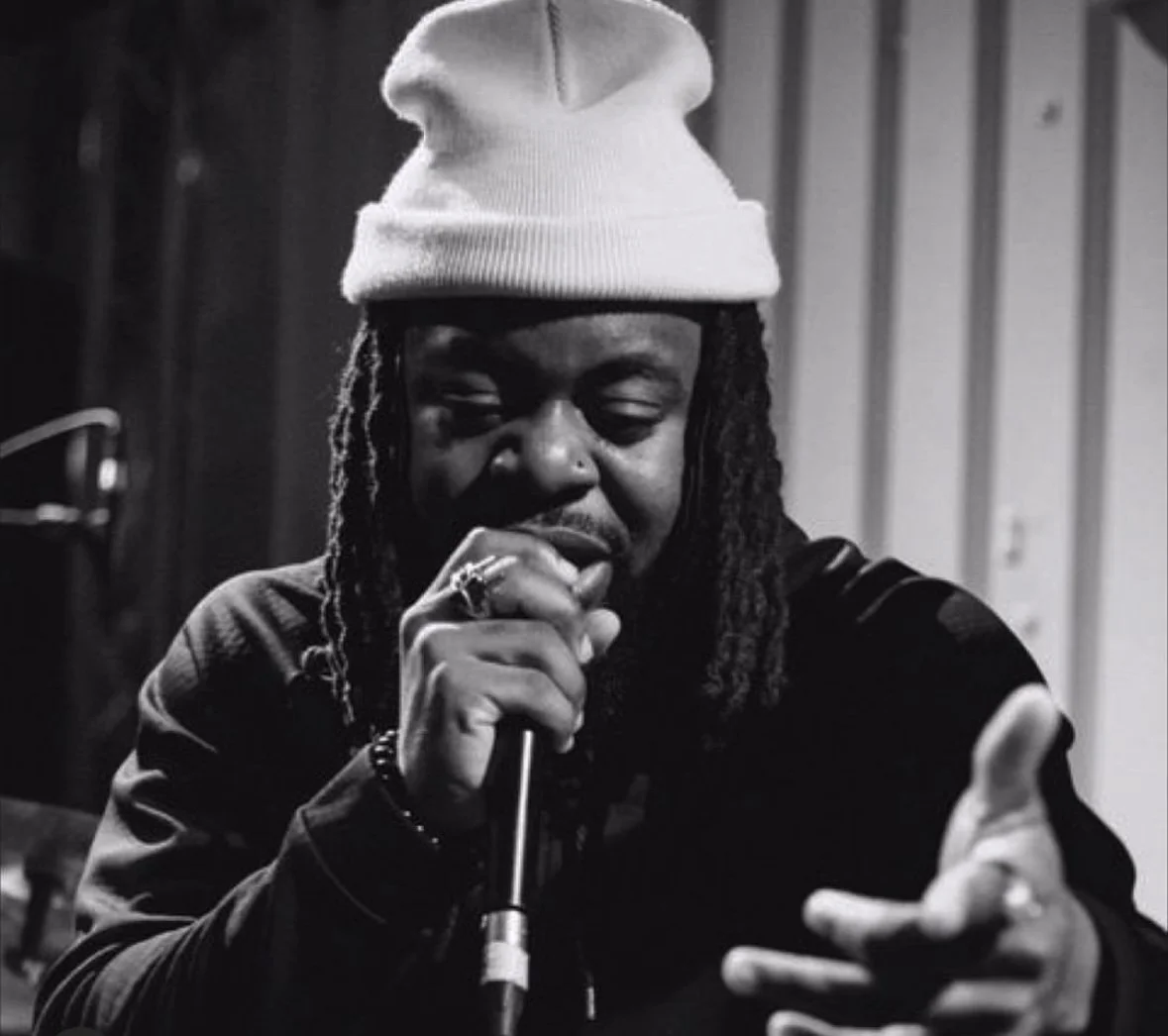 A person with dreadlocks wearing a white beanie, singing or speaking into a microphone in a recording studio, black and white photograph.