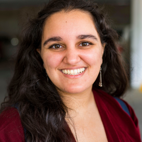 A young woman with dark curly hair, smiling, wearing a maroon top and silver earrings.