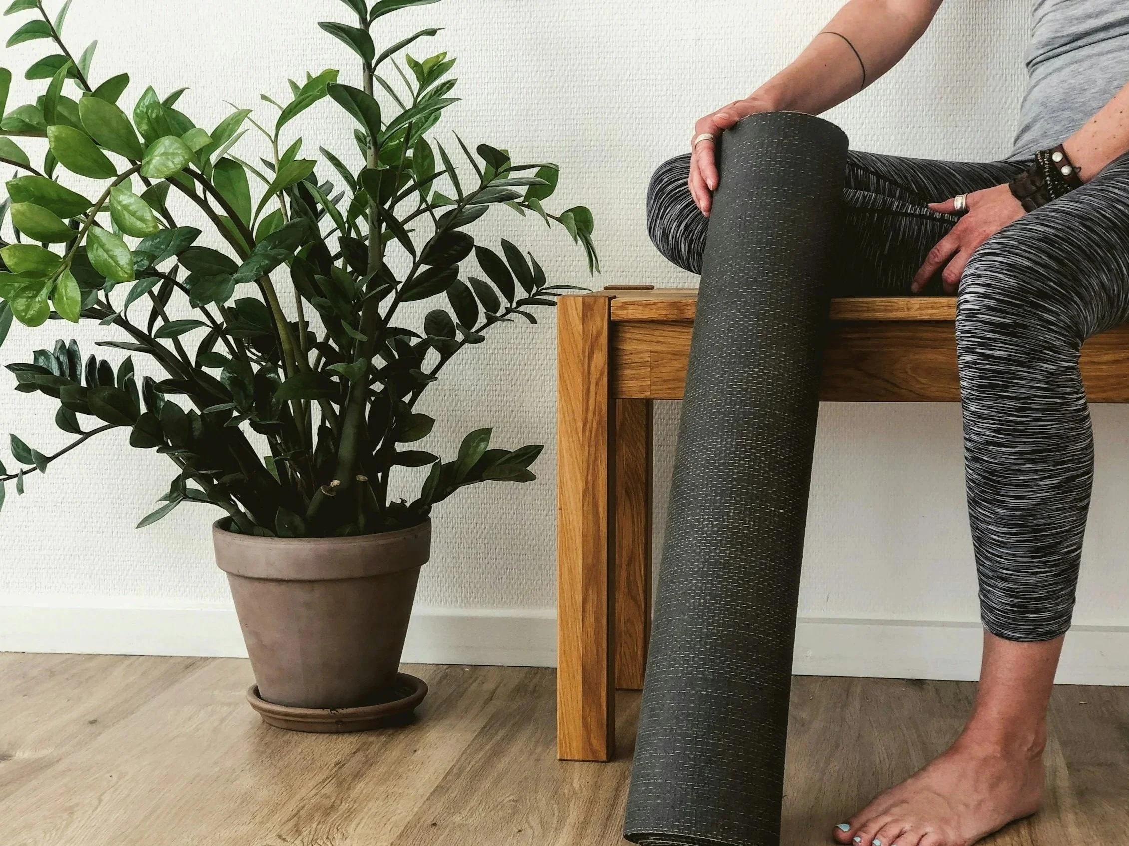 Person sitting on a wooden bench with leggings and a gray t-shirt, holding a black yoga mat while a large green potted plant is nearby.