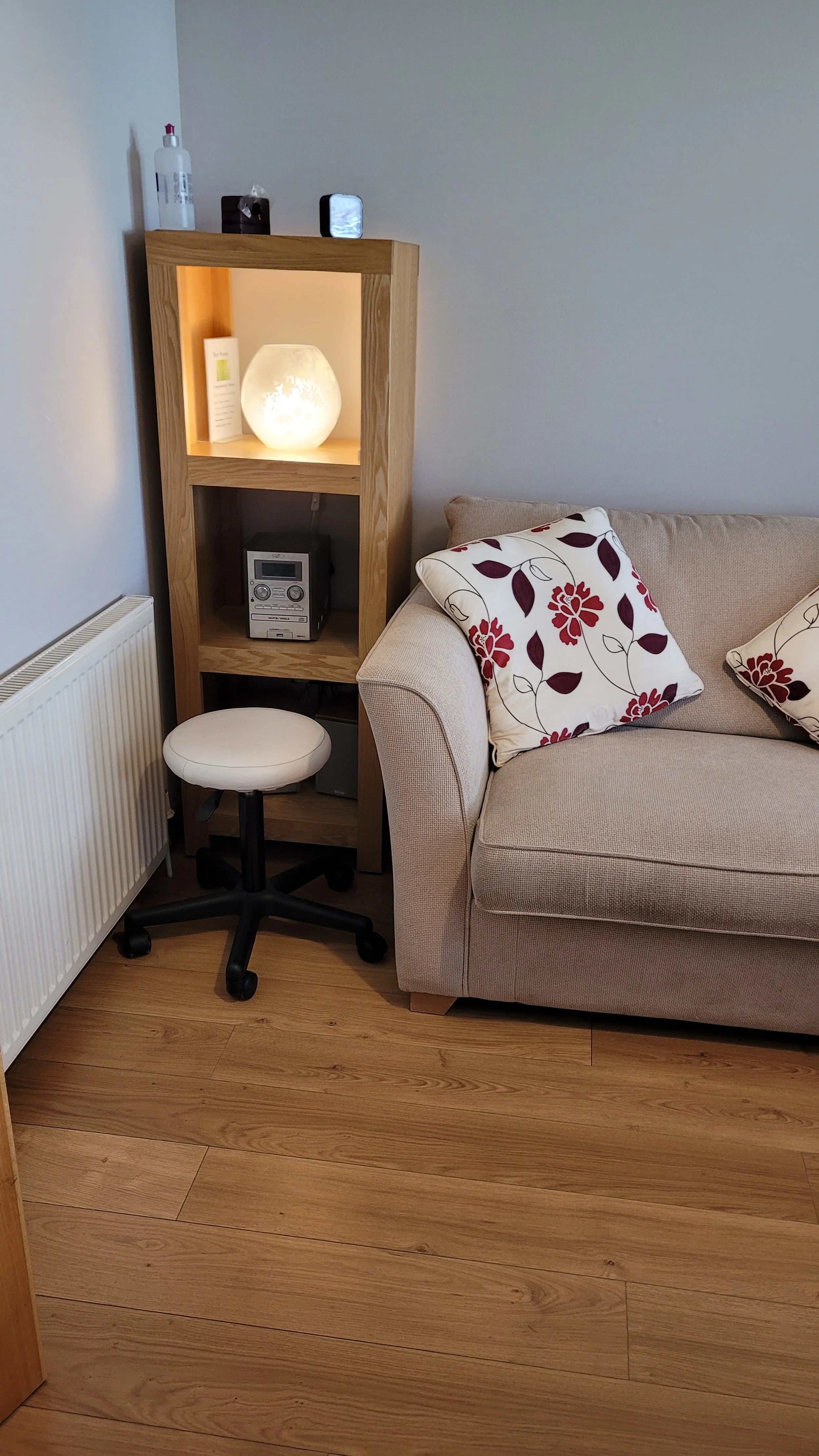 A beige sofa with red and white floral pillows, a wooden shelving unit with a lit decorative lamp, a small stereo, a tissue box, a bottle, and a cellphone, next to a white radiator and a swivel stool on a wooden floor.