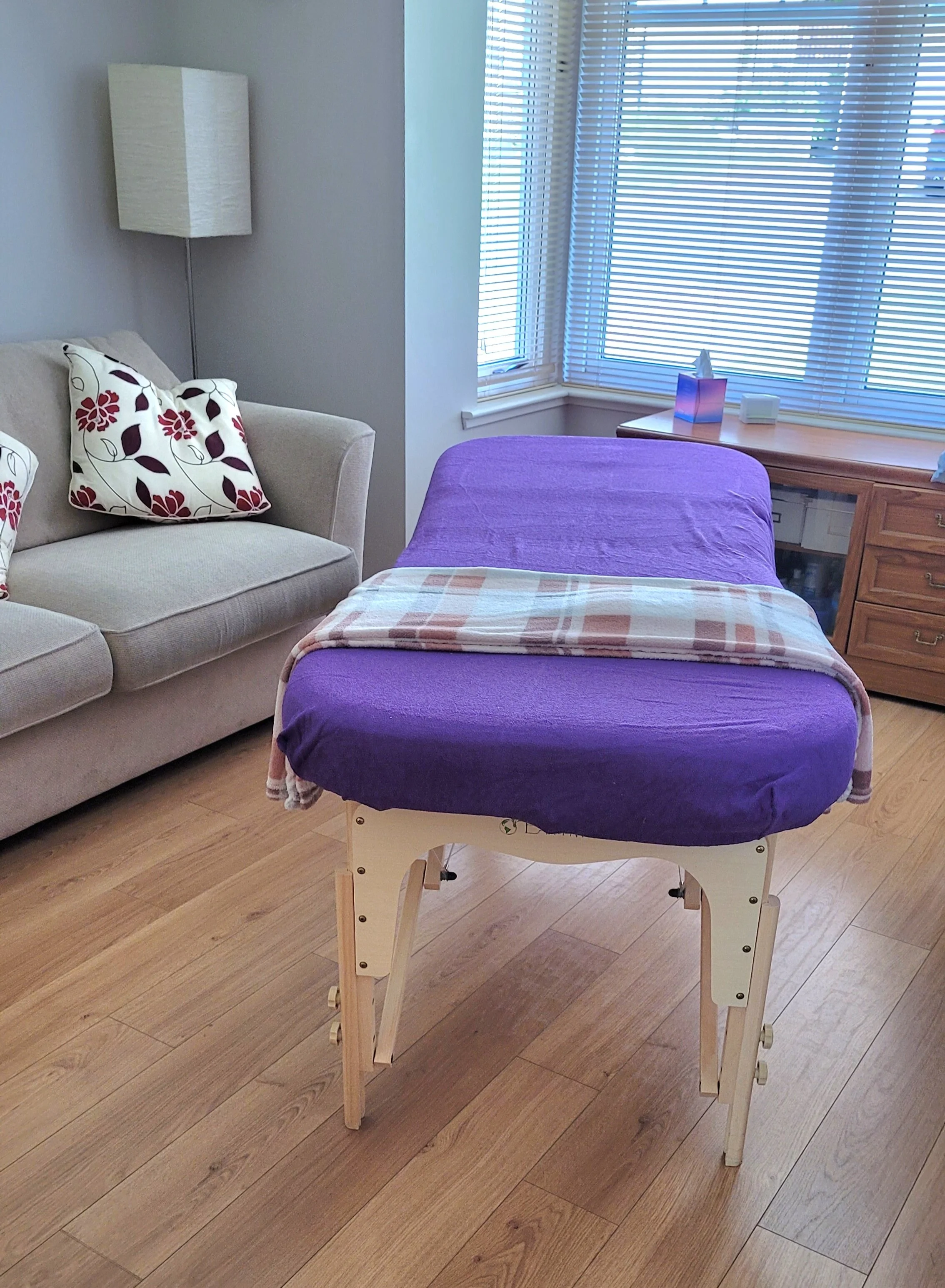 A massage table with a purple cover and a beige and pink striped blanket, set up in a living room with a beige couch, floral pillows, wooden flooring, large windows with blinds, and a wooden side table with tissues and other items.