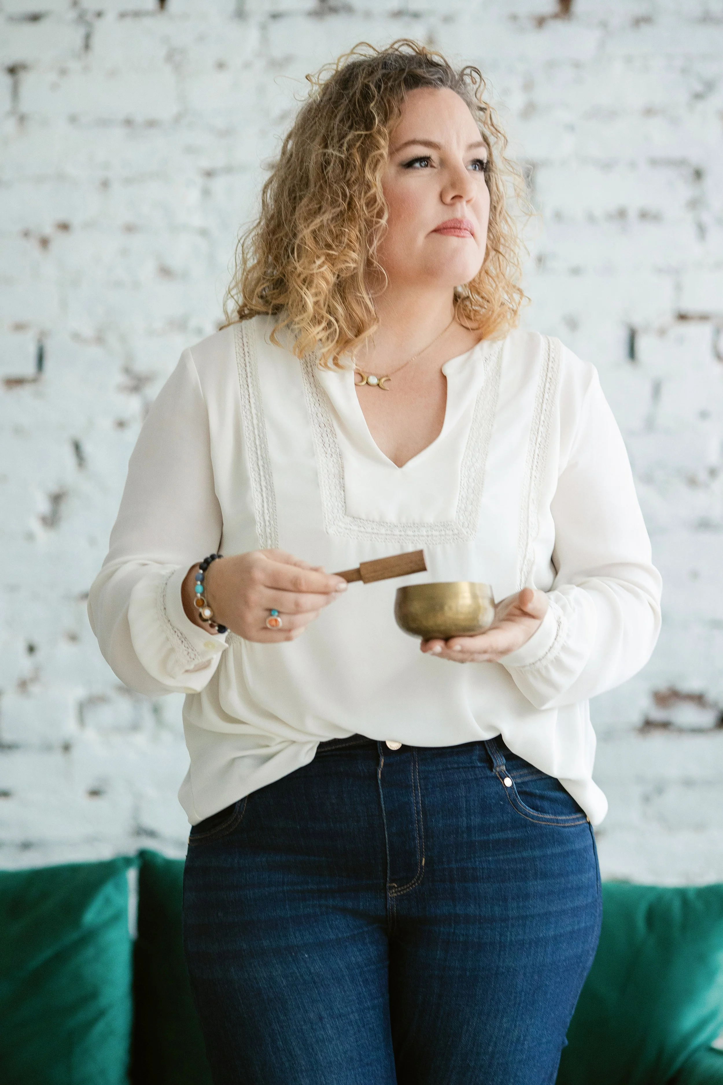 A woman with curly blonde hair holding a singing bowl and a wooden striker, standing near a green sofa against a white brick wall.