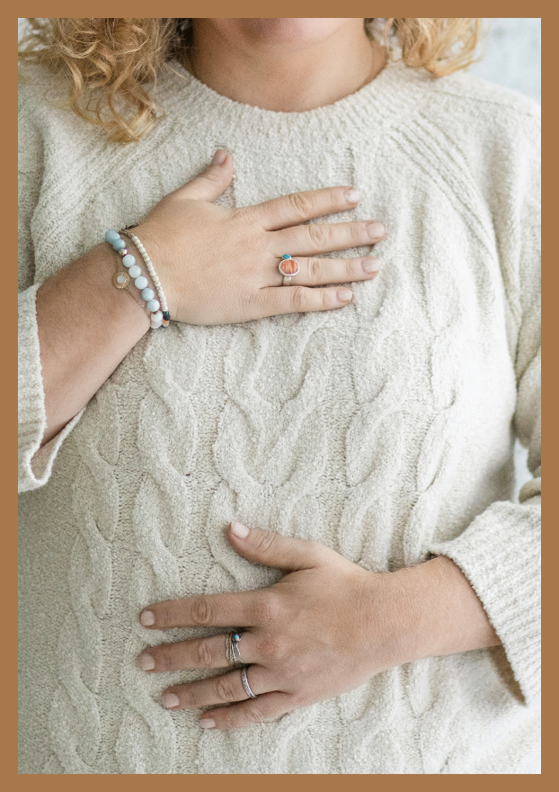 Person with curly hair wearing a cream-colored cable knit sweater, hands on chest and belly, wearing rings and bracelets.