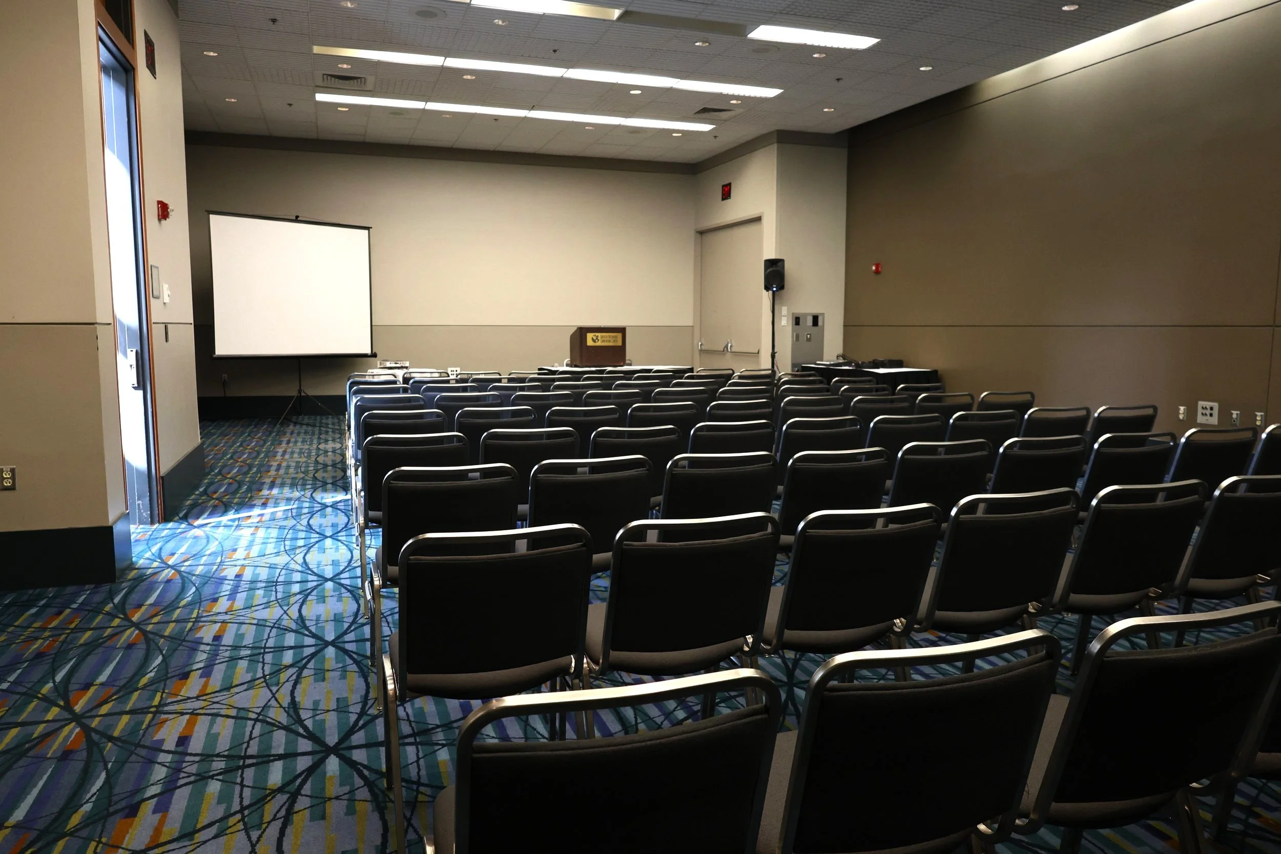 Empty conference room with rows of black chairs, a projector screen, a podium, and audio equipment.