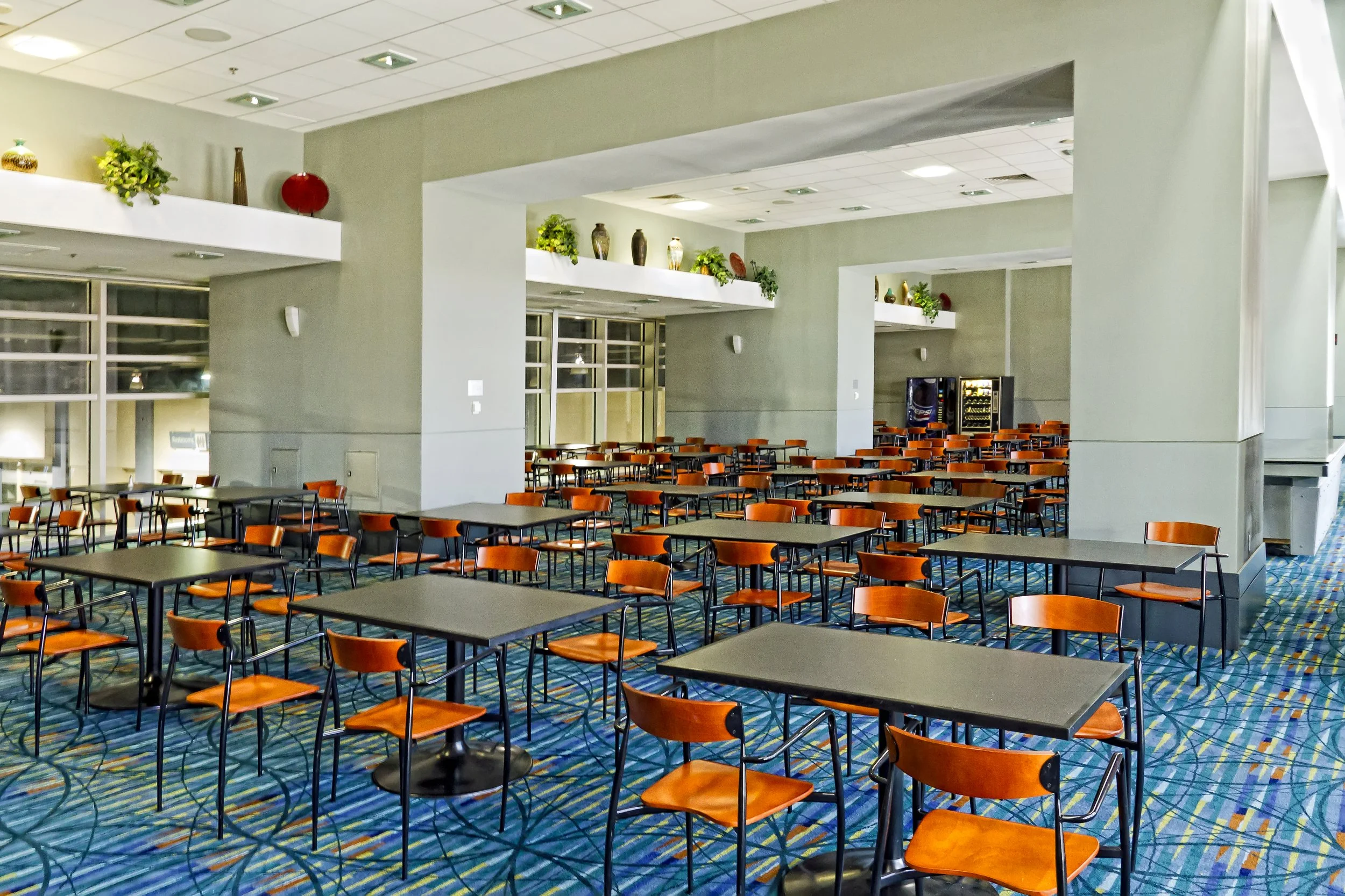 Empty dining area with multiple tables and orange chairs, vending machines in the background, and decorative items on ledges, under a ceiling with recessed lighting.