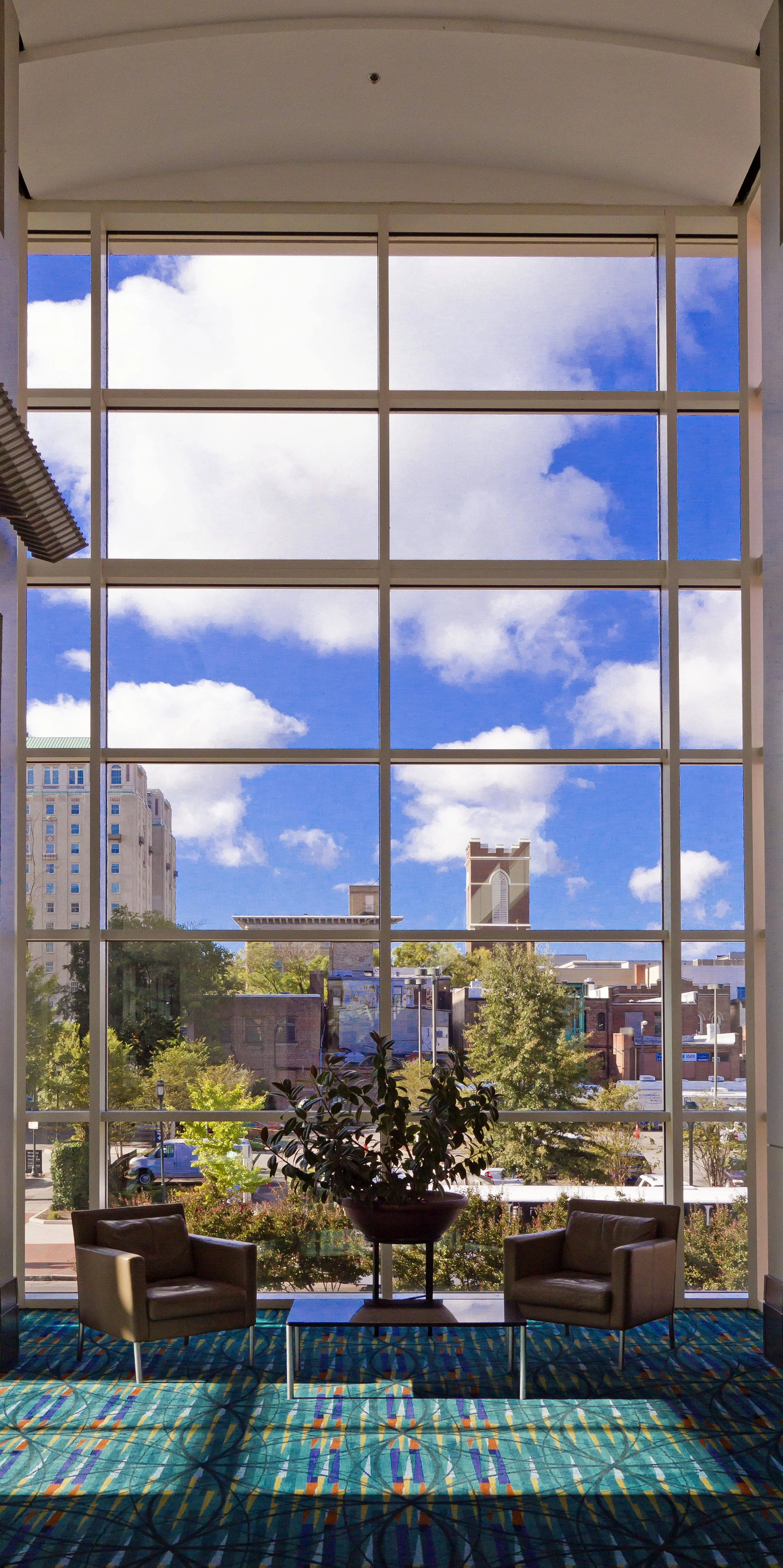 Interior view of a room with large floor-to-ceiling windows looking out onto a cityscape with trees, buildings, and a blue sky with clouds. There are two beige armchairs and a table with a potted plant inside the room, and a colorful patterned carpet