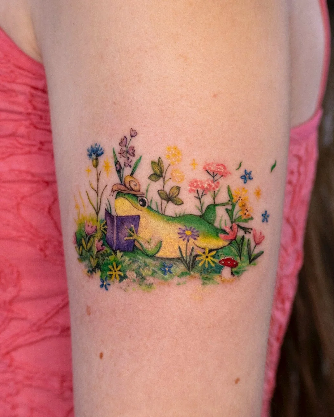 Cute illustrative tattoo design featuring a charming frog sitting among delicate flowers while reading a book. Whimsical nature-inspired artwork with playful details, expressive linework, colorful watercolour feeling and a cosy, storybook feel.