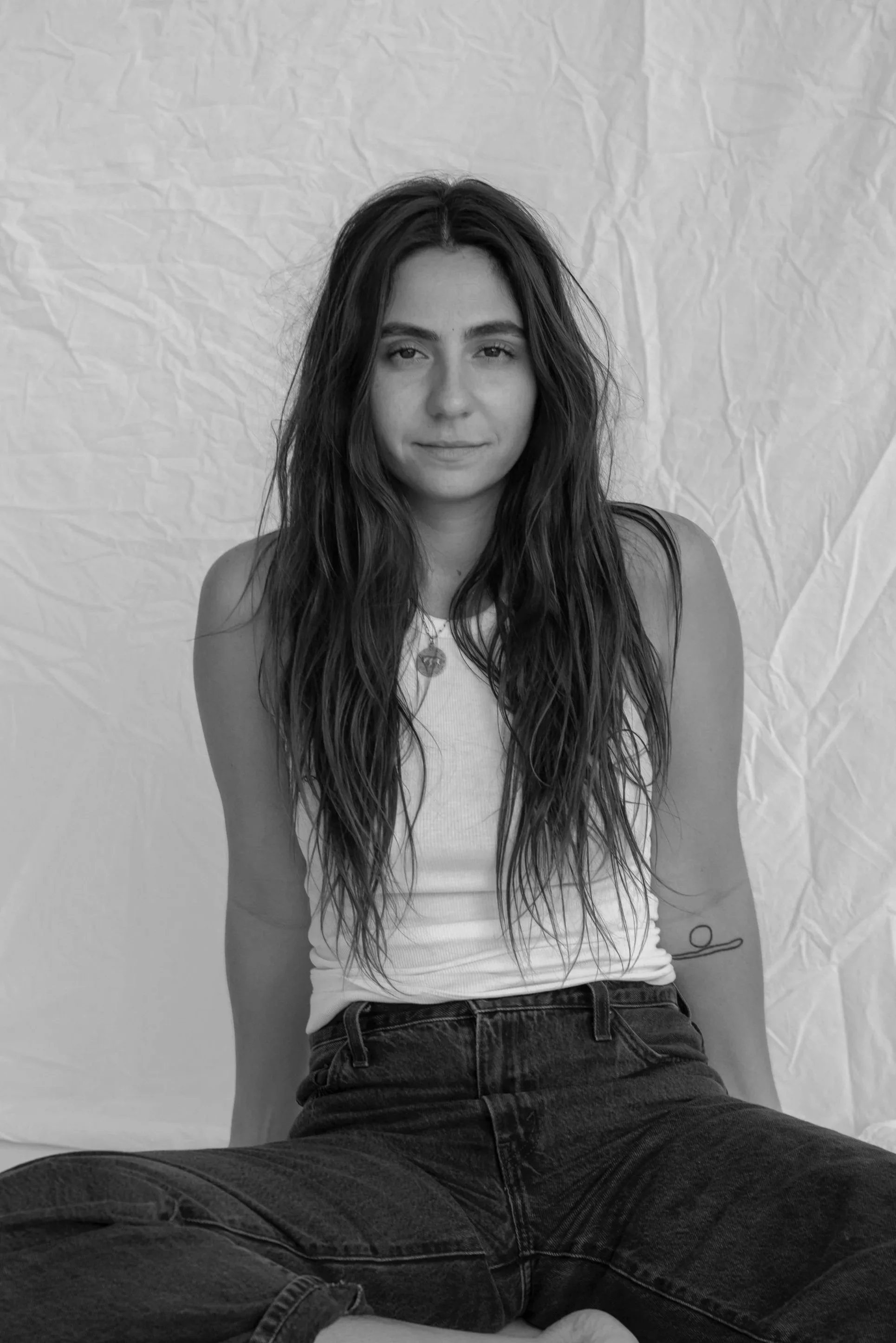 A young woman with long wavy hair sitting against a plain, crumpled background, wearing a sleeveless top and jeans, looking at the camera. Black and white photo.