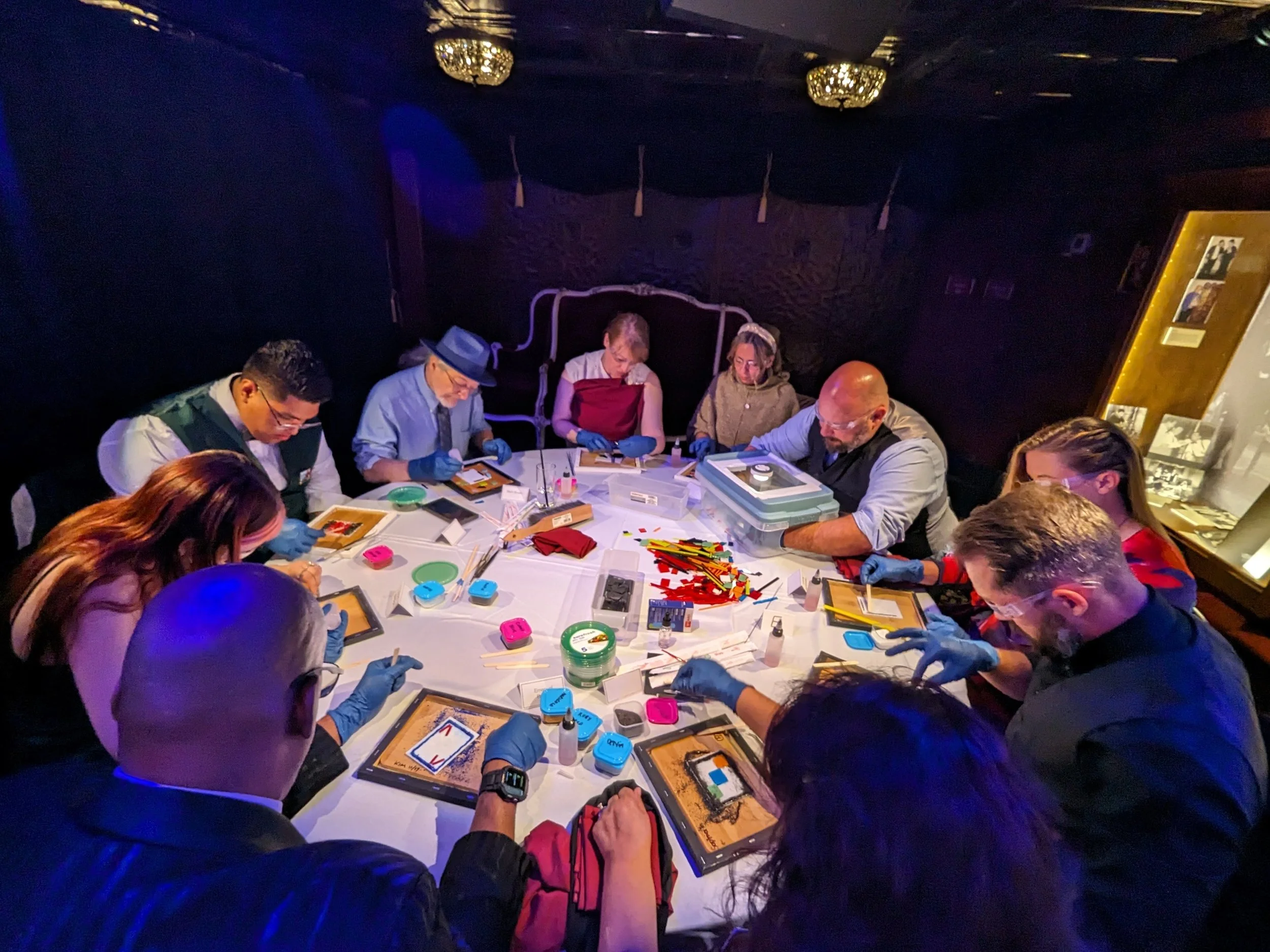 A group of nine people sitting around a table engaged in a collaborative activity, possibly an escape room or puzzle-solving game. They are using tablets or framed screens, and all are wearing blue gloves. The table has various colorful game pieces, 