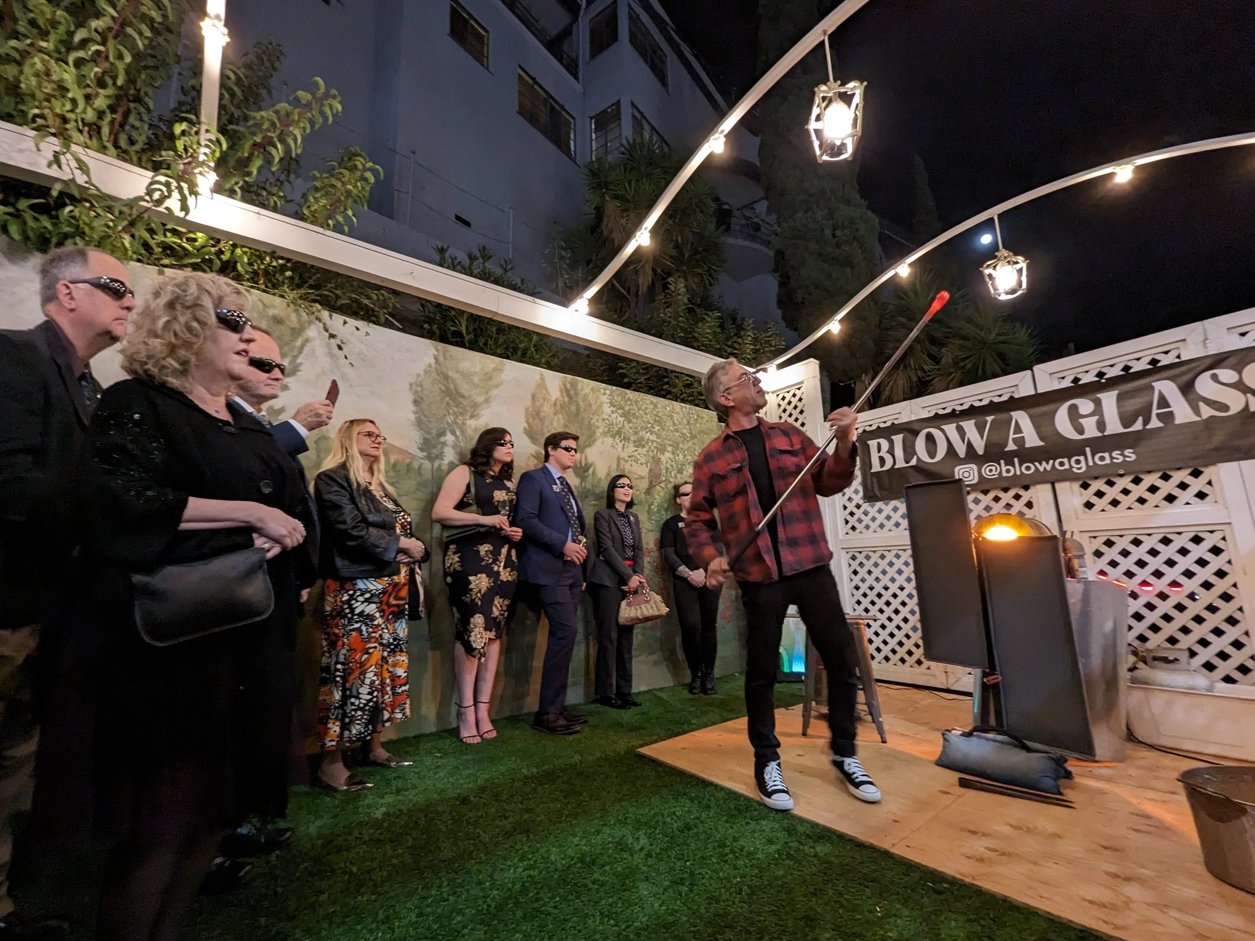 A group of people wearing sunglasses and formal attire watching a man in a red plaid jacket blow glass with a long metal tool, under string lights at night. There is a banner reading 'BLOW A GLASS' in the background.
