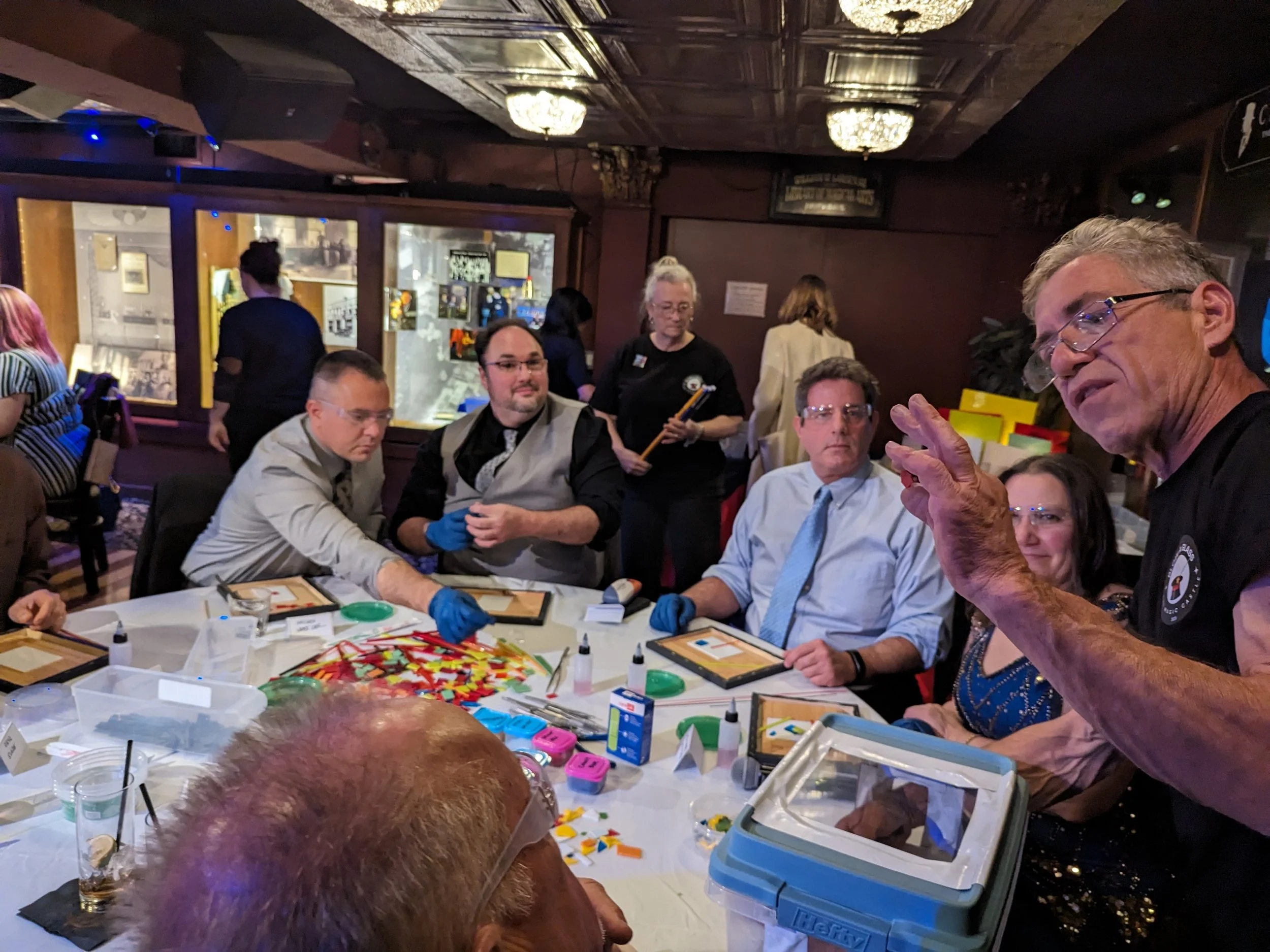 A group of people gathering around a table with game pieces, cards, and droppers, as a man explains something to the group in a dimly lit room with chandeliers.