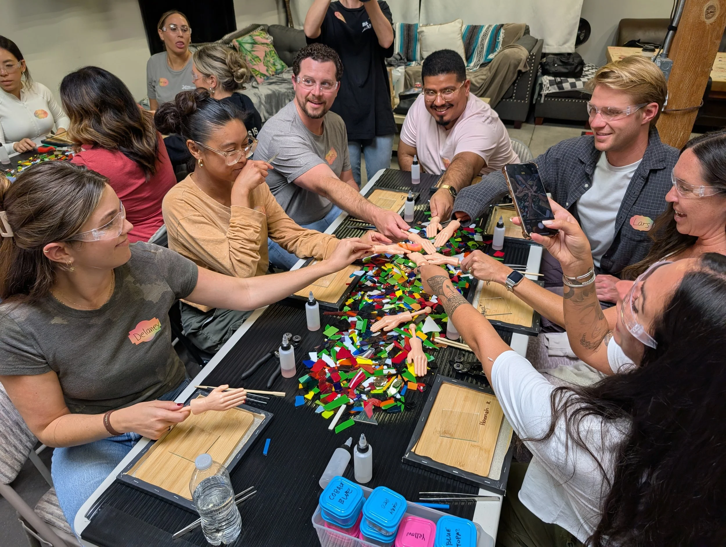 People gathered around a table participating in a mosaic art activity, using colorful tiles and small figurines, with some members taking photos and sharing the experience. Teambuilding event for groups.